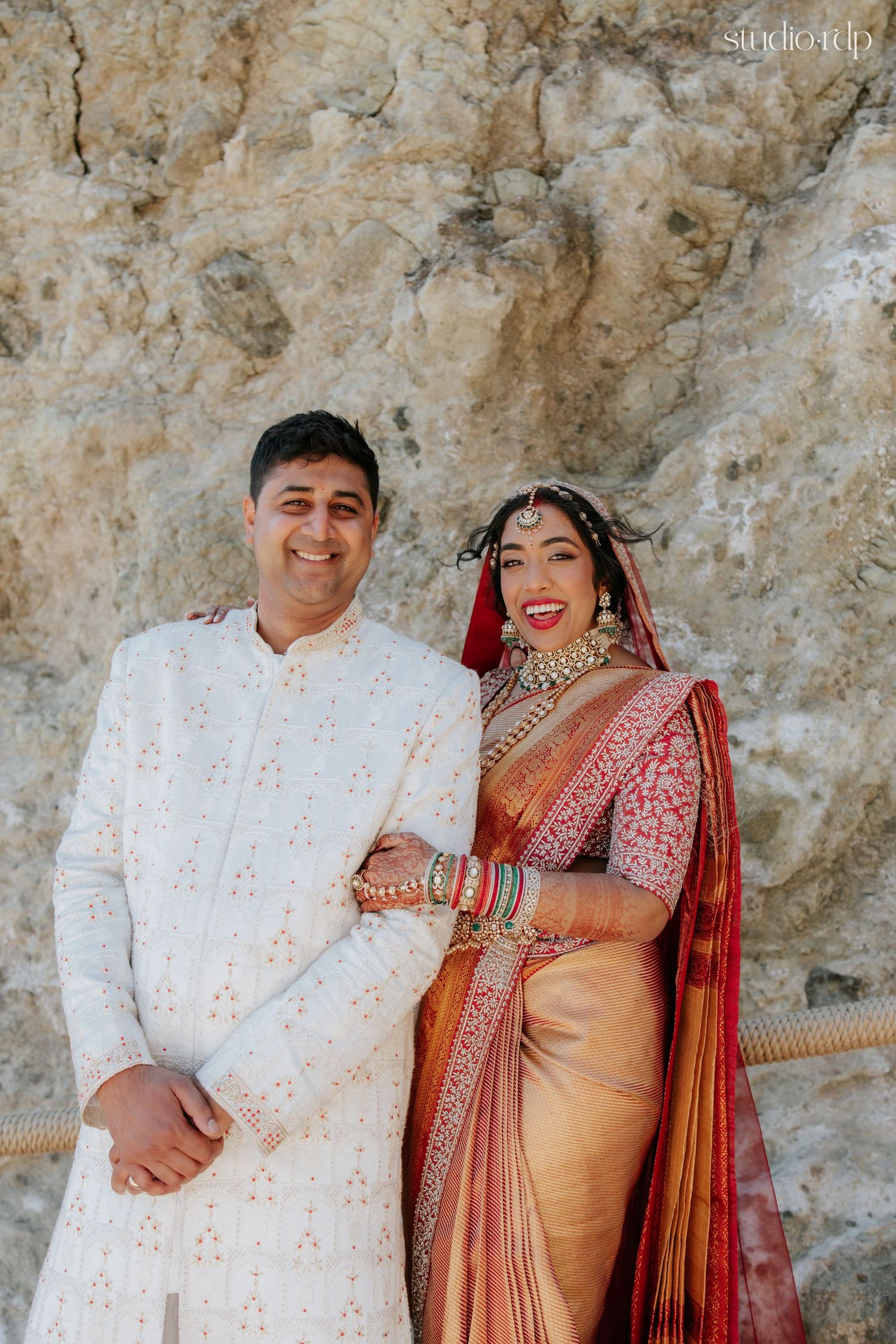 Indian couple in traditional wedding attire, groom in white sherwani and bride in red and gold saree with bridal jewelry.