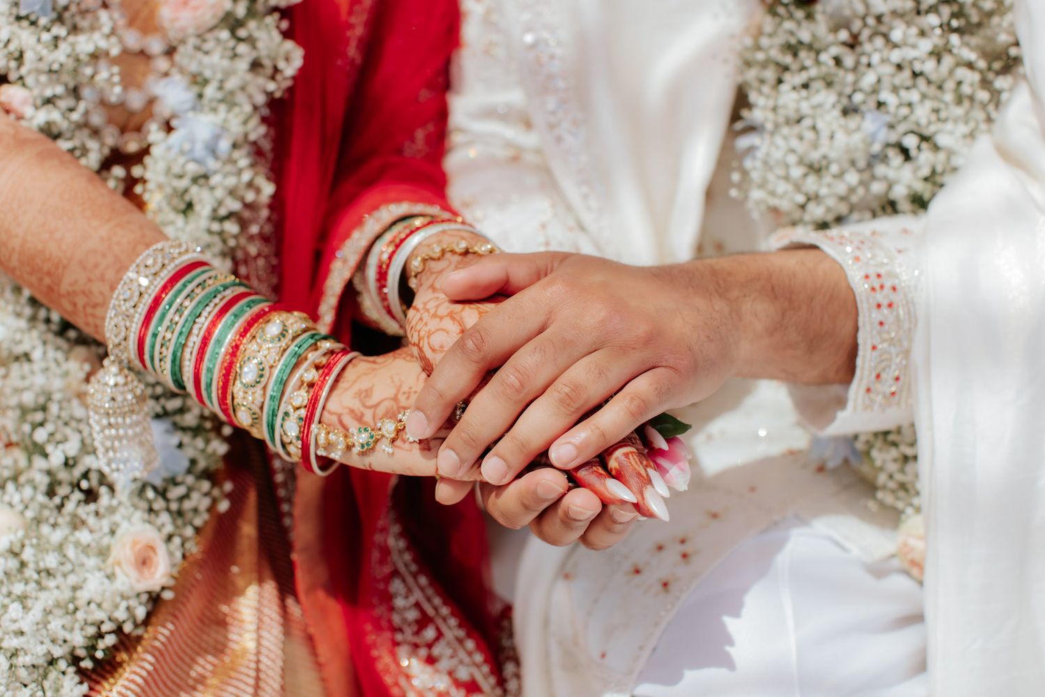 Close-up of Indian bride and groom holding hands, bride wearing red saree, mehndi, and colorful bangles at wedding.