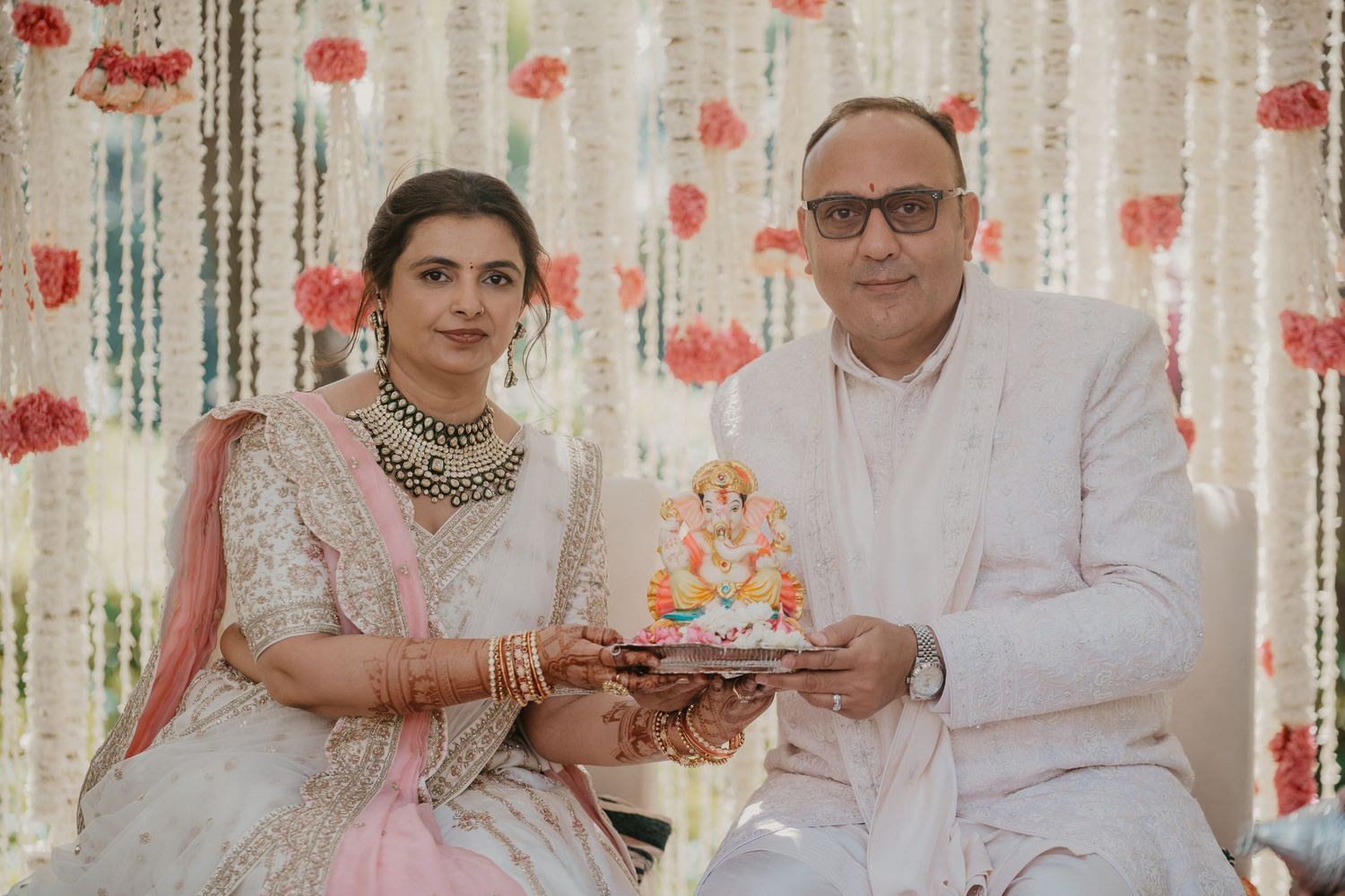 A couple in traditional Indian attire pose together at a ceremony with a background of pink floral decorations.