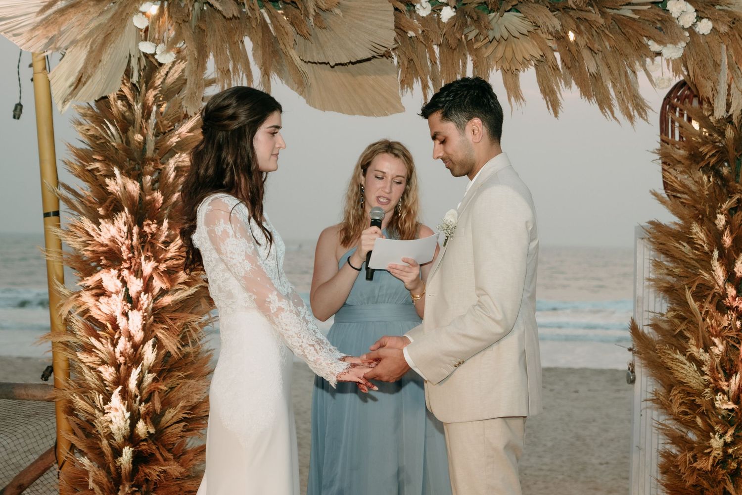 A wedding ceremony on the beach at sunset under a pampas grass arch with decorative lighting.