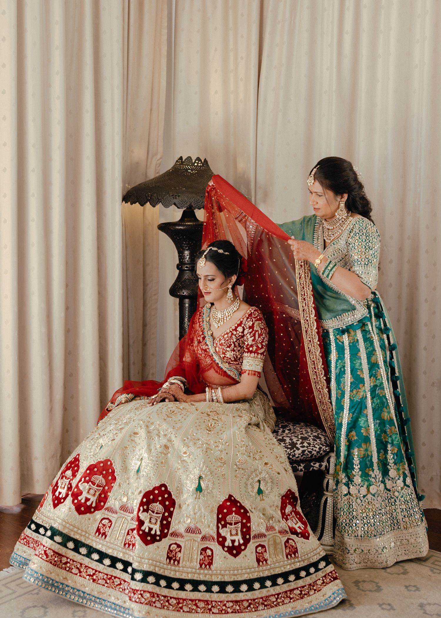 Traditional Indian bride in red and white lehenga being helped with her dupatta against beige curtain backdrop.