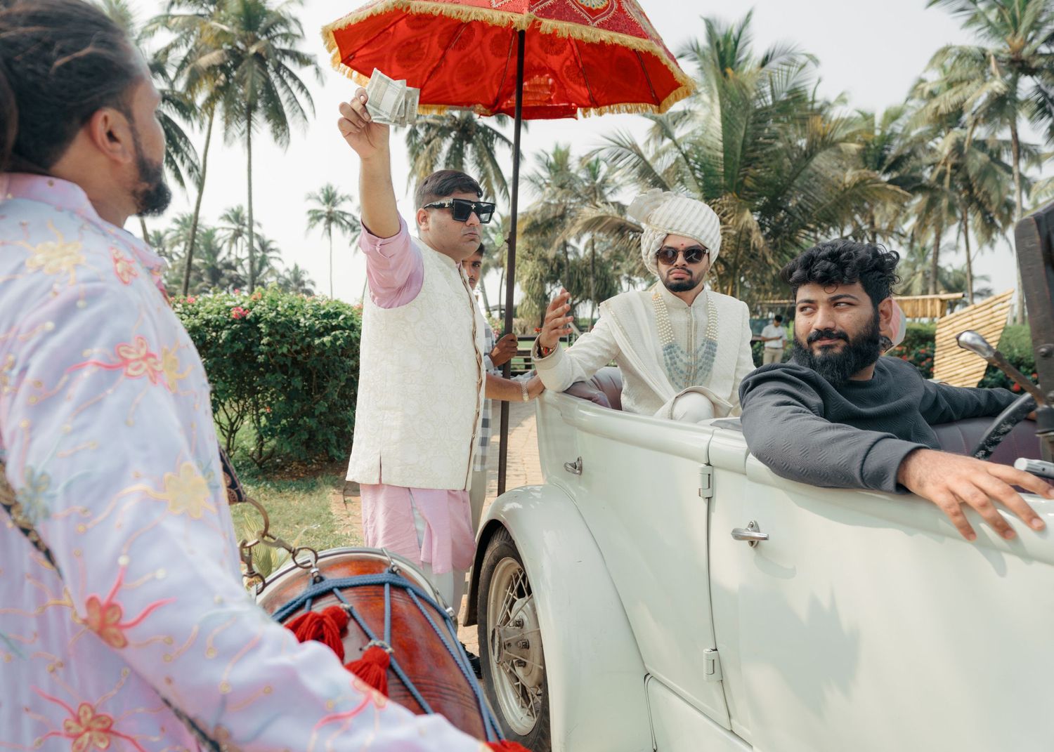 Three people enjoy a ride in a vintage white convertible car under a red umbrella surrounded by palm trees.