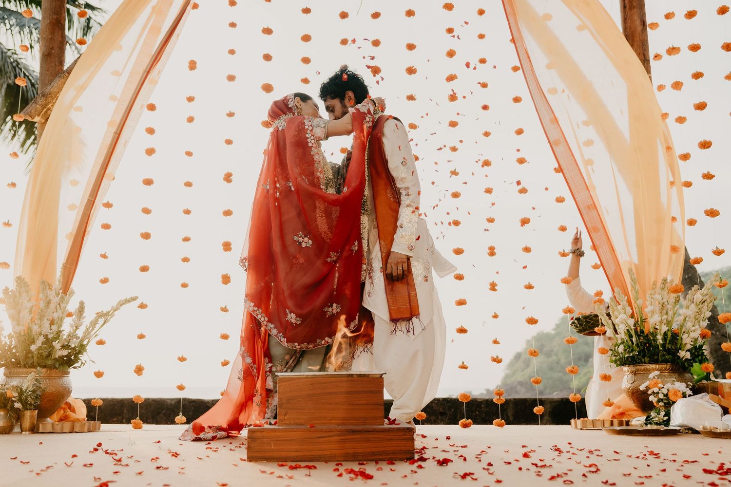 Indian bride and groom share a kiss during traditional wedding ceremony with floral backdrop and rose petals.