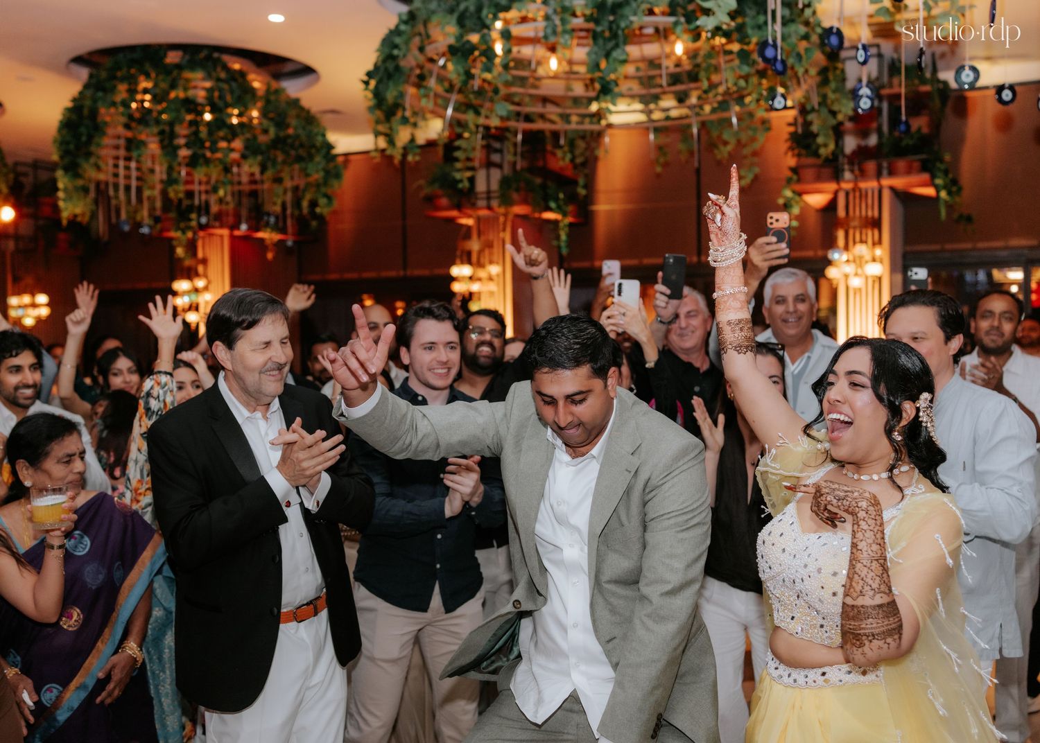 Guests celebrate joyfully at an Indian wedding reception with colorful floral decor and raised hands.