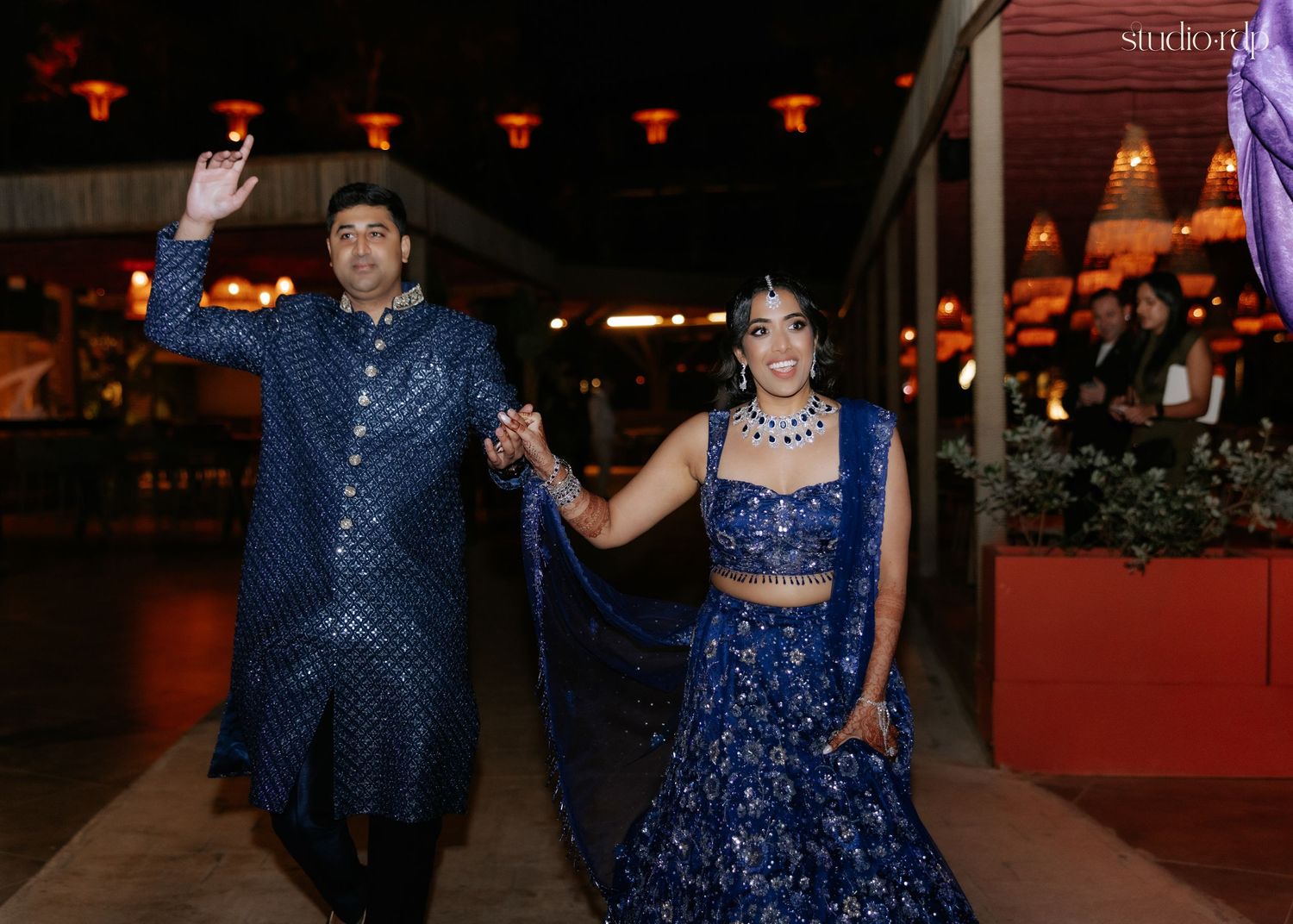 Couple in matching royal blue Indian wedding attire walking hand in hand at an elegant evening celebration.