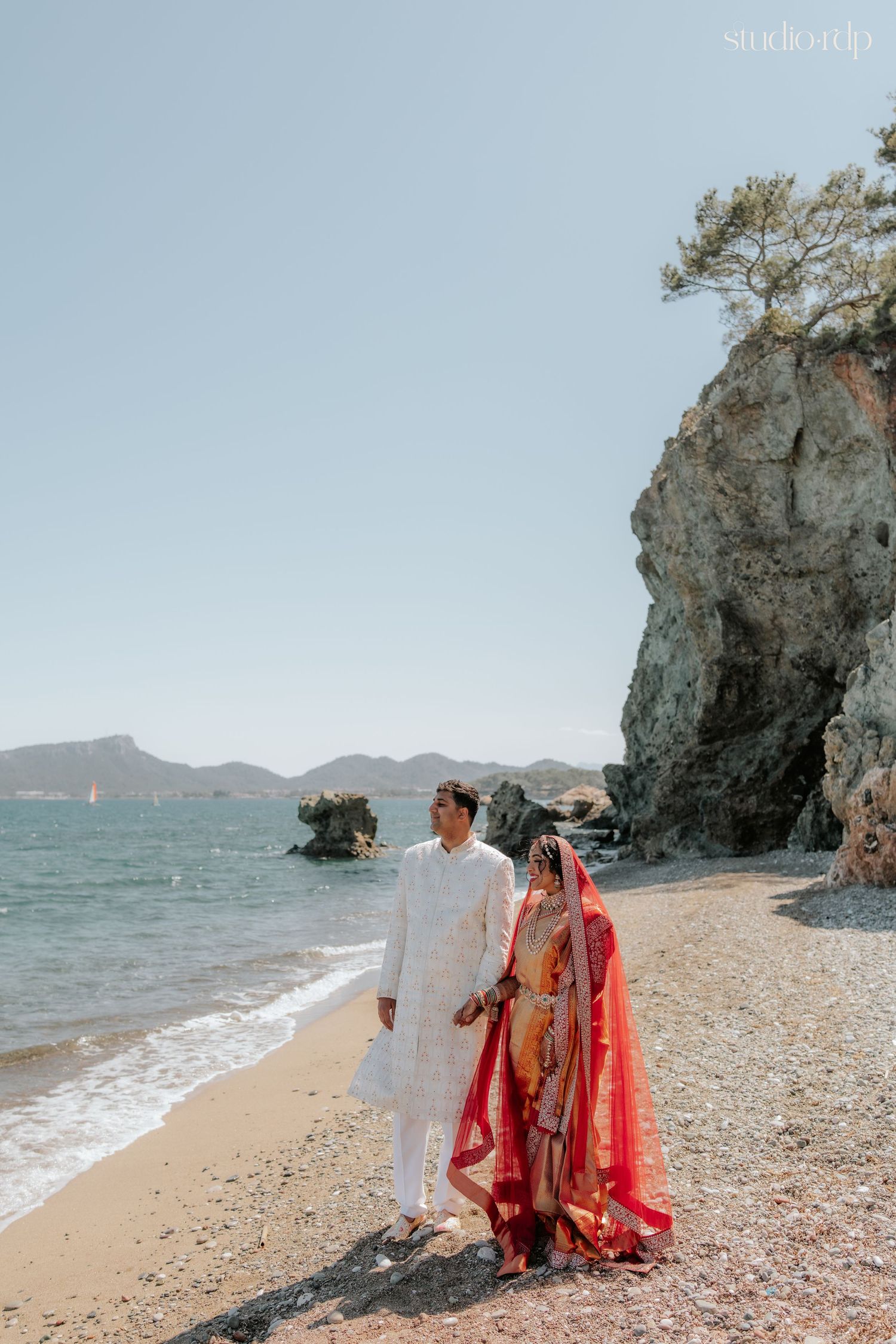 Indian bride in red saree and groom in white sherwani pose on a scenic beach with rocky cliffs.