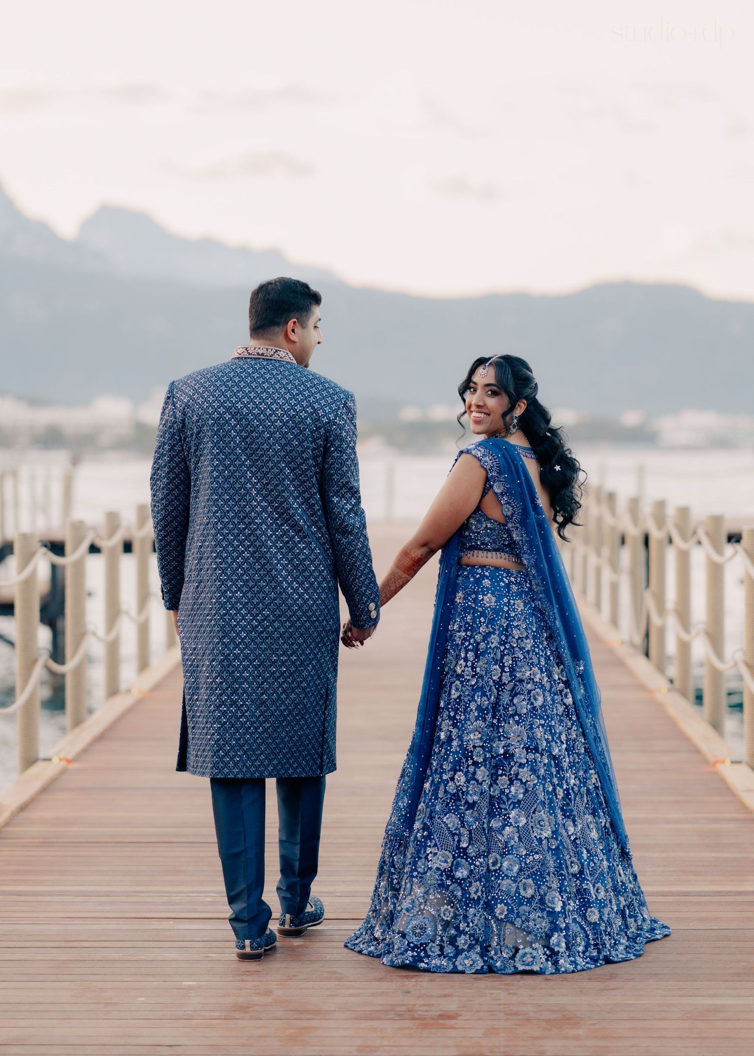 Indian couple in matching blue outfits holding hands on a wooden pier with mountain backdrop.