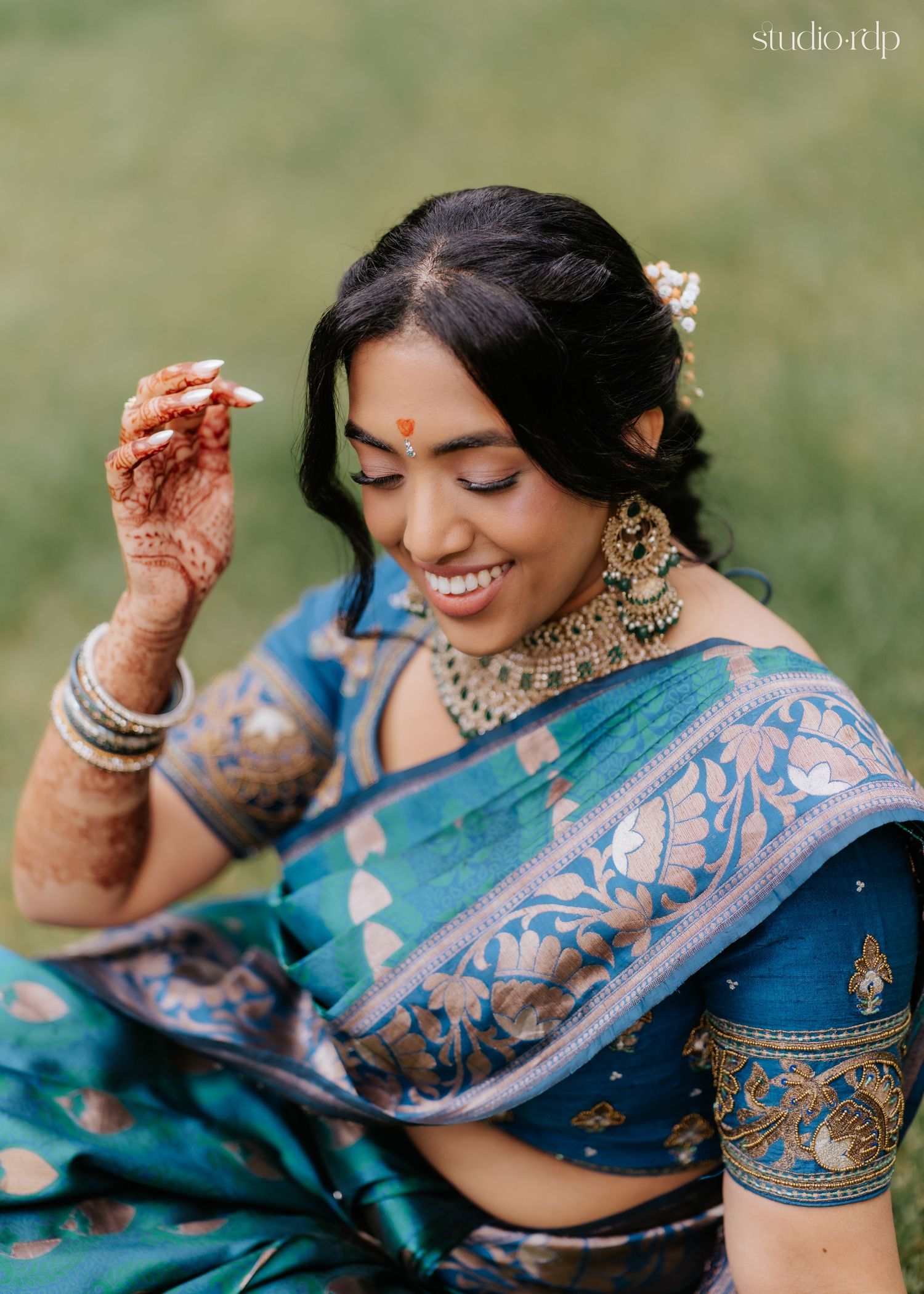 Smiling Indian bride in teal blue silk saree with gold jewelry, mehndi hands, and floral hair accessories outdoors.