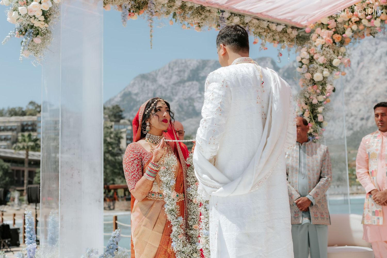 Bride in red saree exchanges garlands with groom in white sherwani during traditional Indian wedding ceremony outdoors.