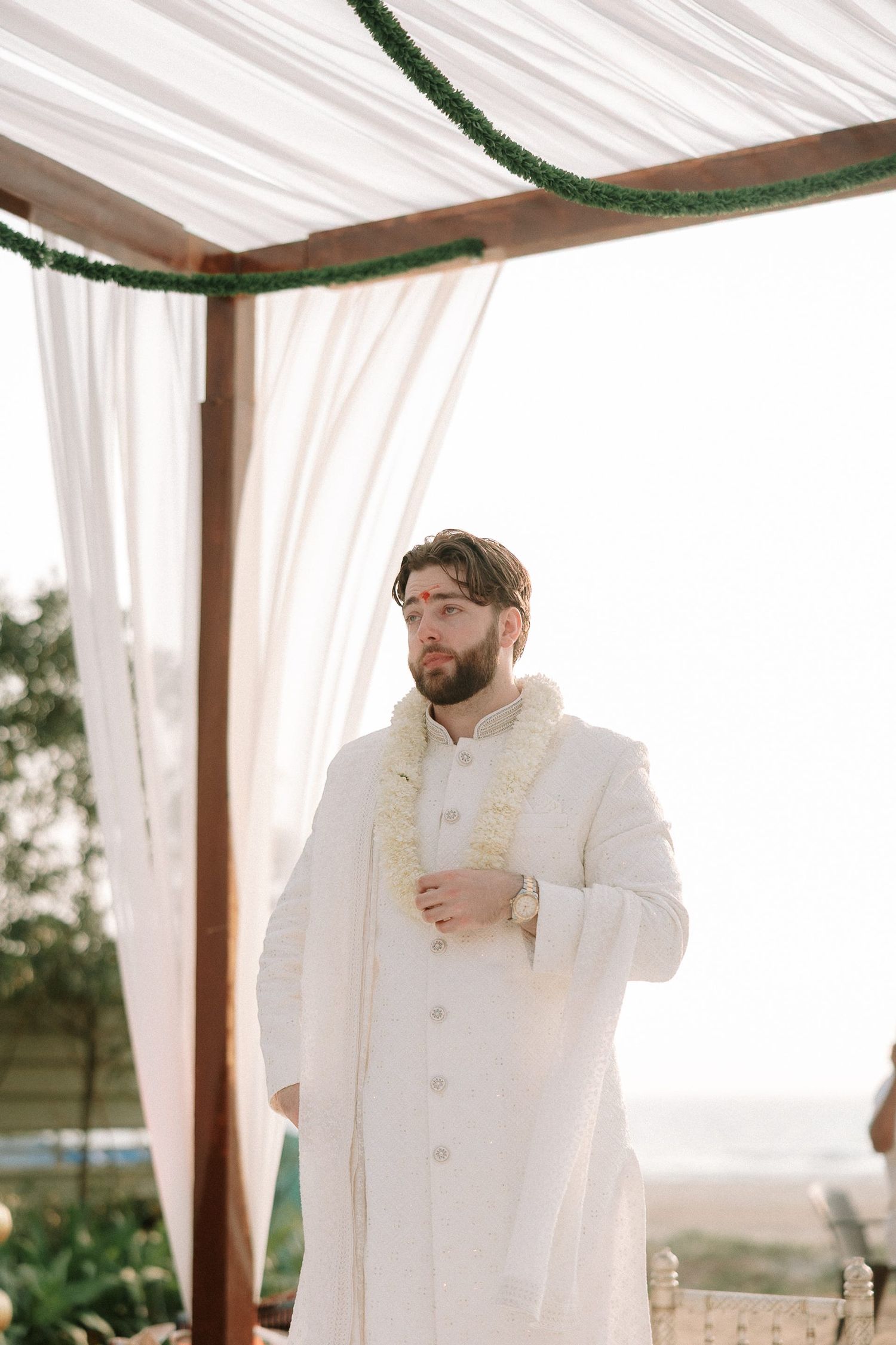 Groom in white sherwani with floral garland stands under decorated mandap canopy at outdoor Indian wedding ceremony.