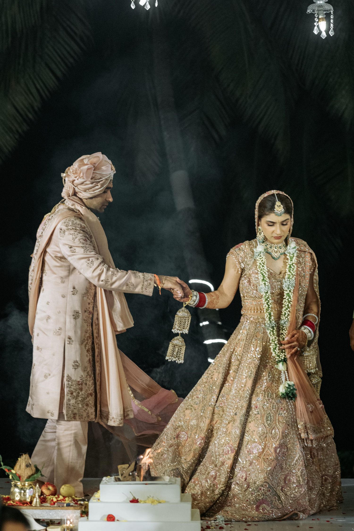 Traditional Indian wedding ceremony with couple in ornate outfits performing ritual under palm fronds.
