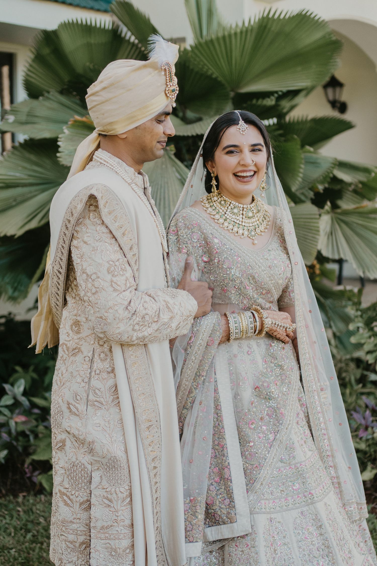 Traditional South Asian wedding couple in matching cream and beige embroidered outfits with palm trees in background.
