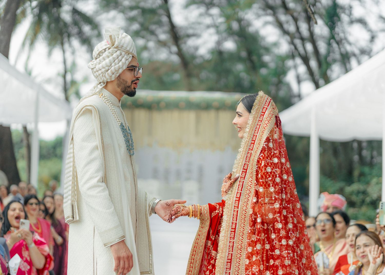 Traditional Indian wedding ceremony moment with couple in elegant cream sherwani and red lehenga attire against palm trees.
