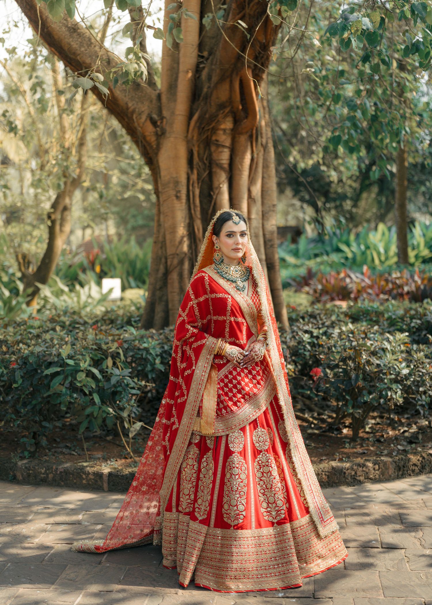 Traditional Indian bride wearing a stunning red and gold embroidered lehenga poses elegantly under trees in an outdoor setting.