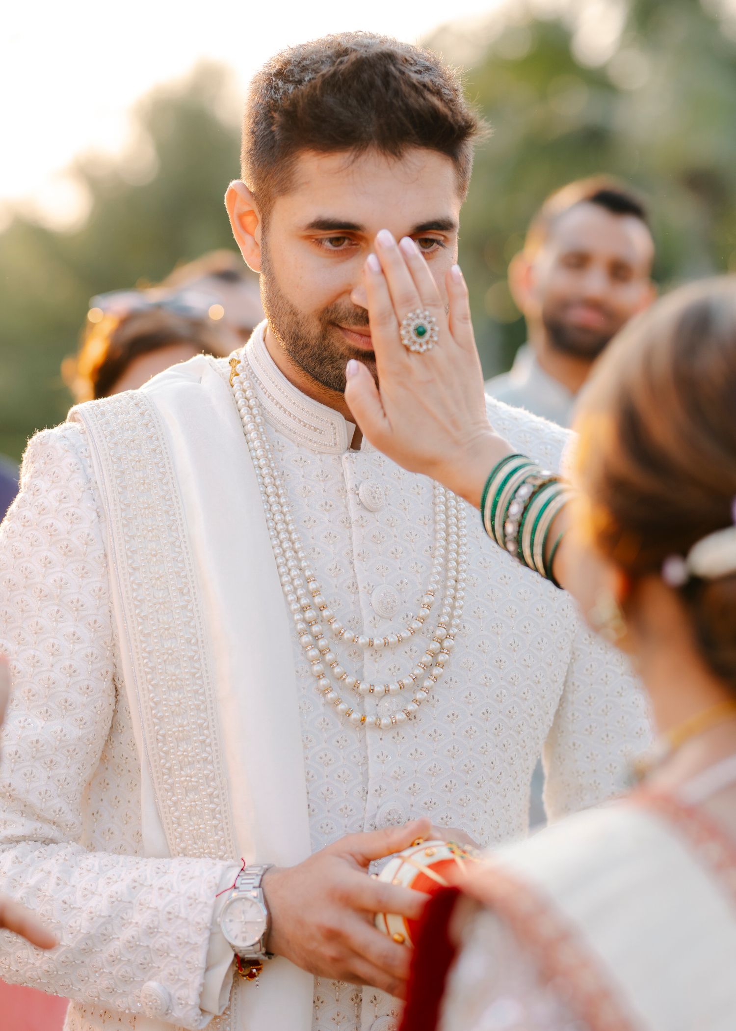 A person in a white embroidered sherwani with pearl necklaces wipes away tears during an emotional wedding ceremony moment.