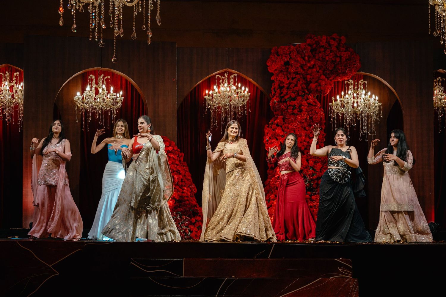 Seven women in elegant gowns perform on a decorated stage with red floral arrangements and chandeliers at a formal event.