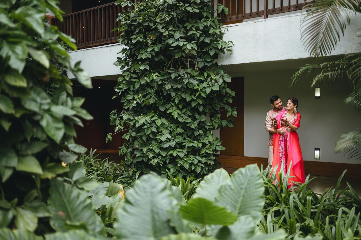Couple in vibrant pink traditional attire standing amidst lush tropical greenery at an elegant garden venue.