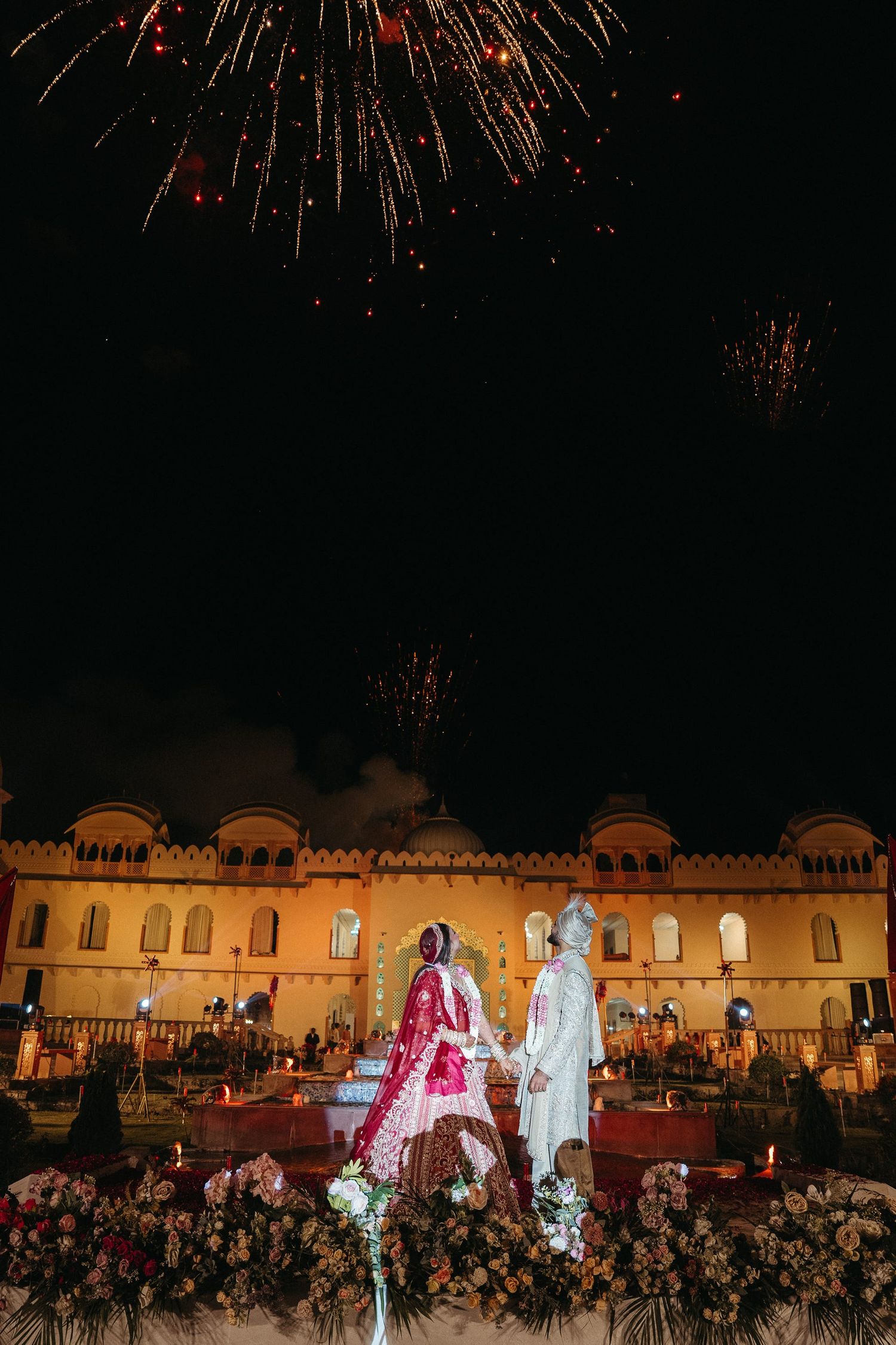 Indian wedding couple in traditional attire hold hands before illuminated palace with colorful fireworks at night.