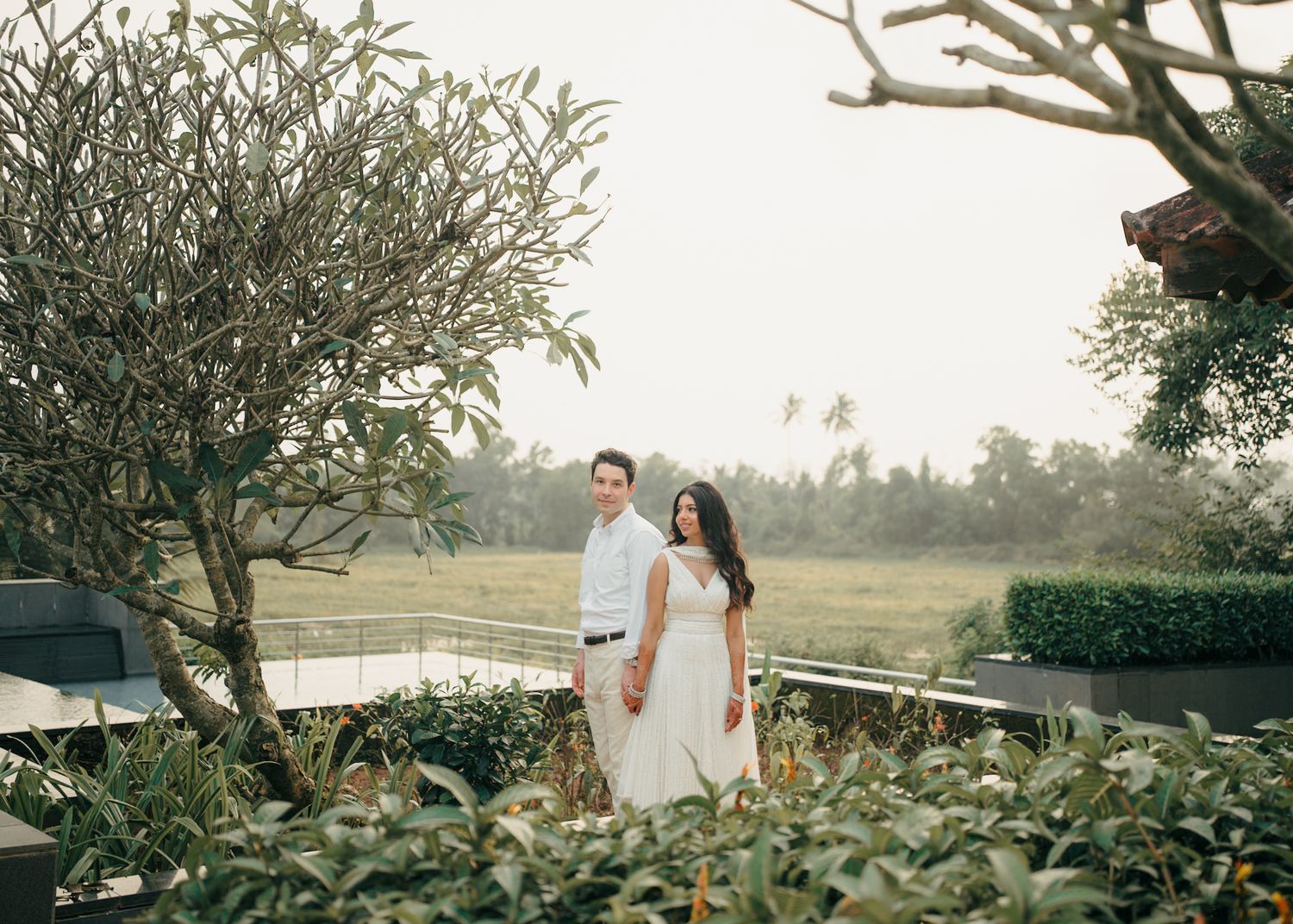 A romantic outdoor photoshoot in a lush garden setting with palm trees and natural greenery at sunset.