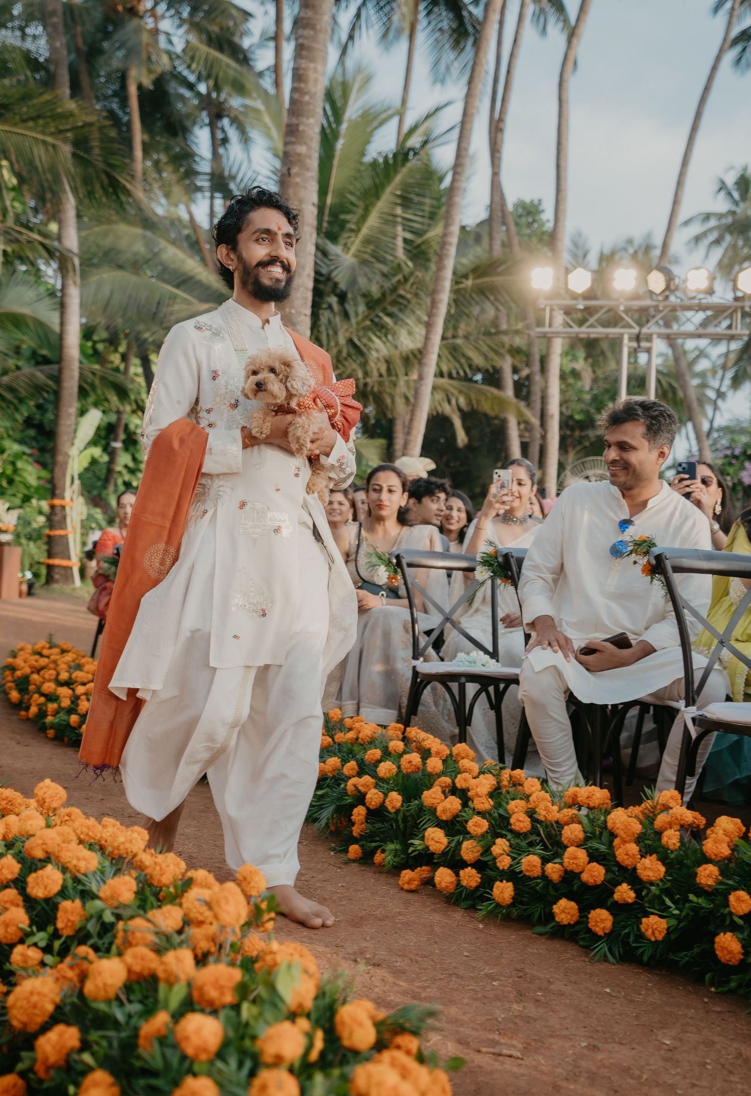 Groom in white kurta carries a dog down the aisle lined with marigolds at an outdoor Indian wedding.