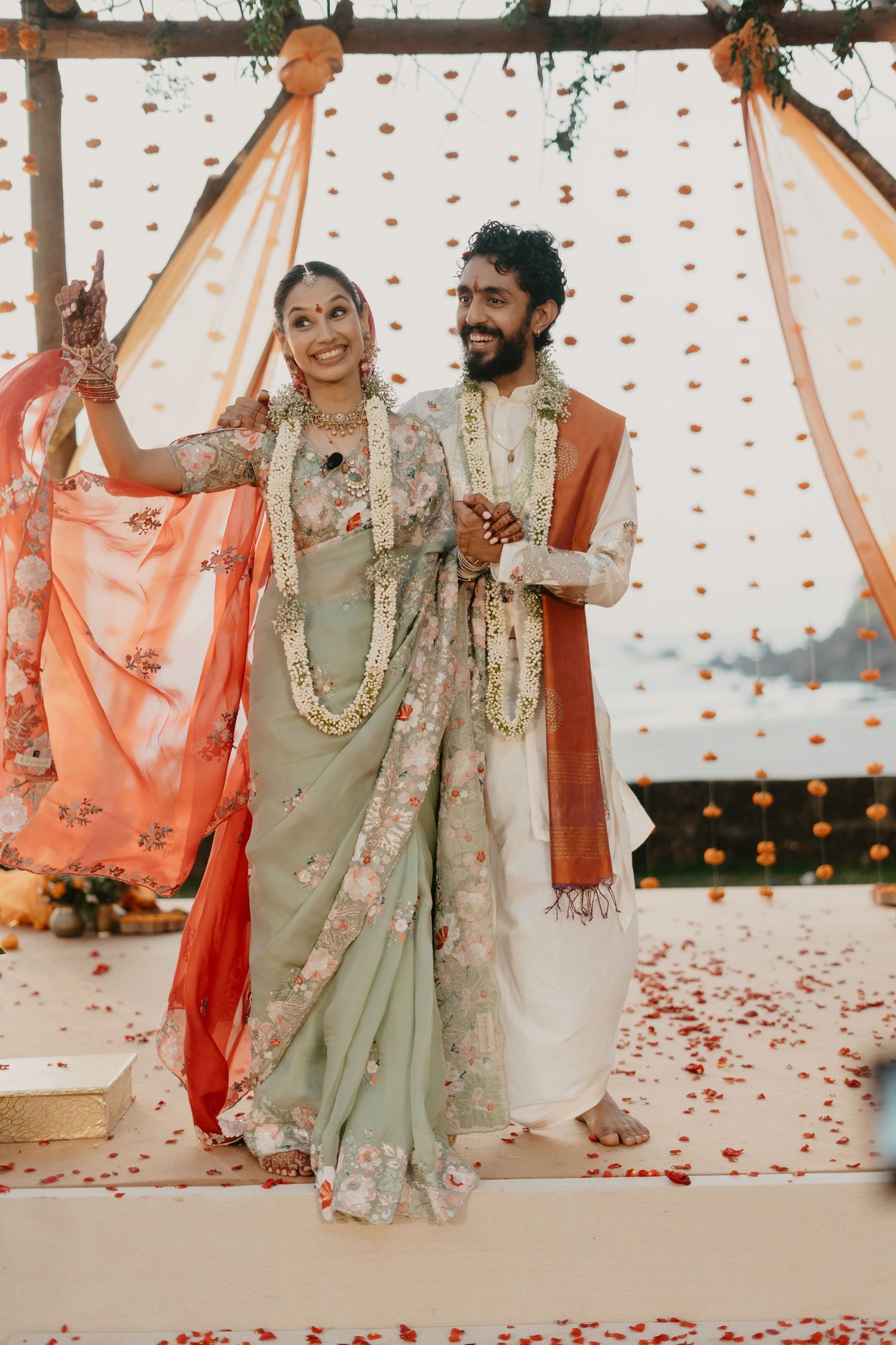 Indian bride in sage green floral saree and groom in white sherwani celebrate at traditional wedding ceremony.