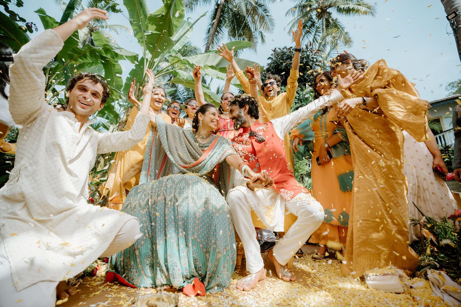 Joyful Indian wedding celebration with guests in colorful traditional attire dancing outdoors amid falling flower petals.