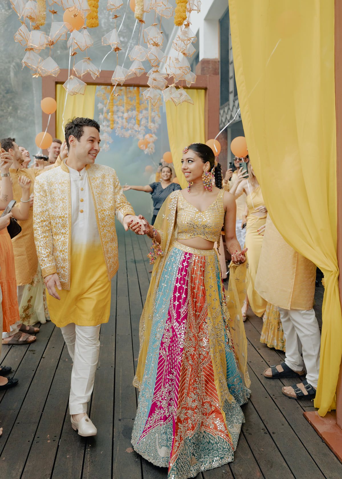 Couple in vibrant Indian wedding attire walk hand-in-hand under floral decorations at a colorful Haldi ceremony.