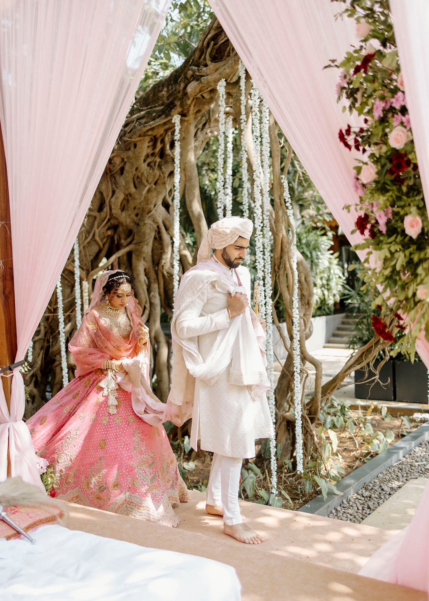 Indian bride in pink lehenga and groom in white sherwani walking together under floral mandap decorations.
