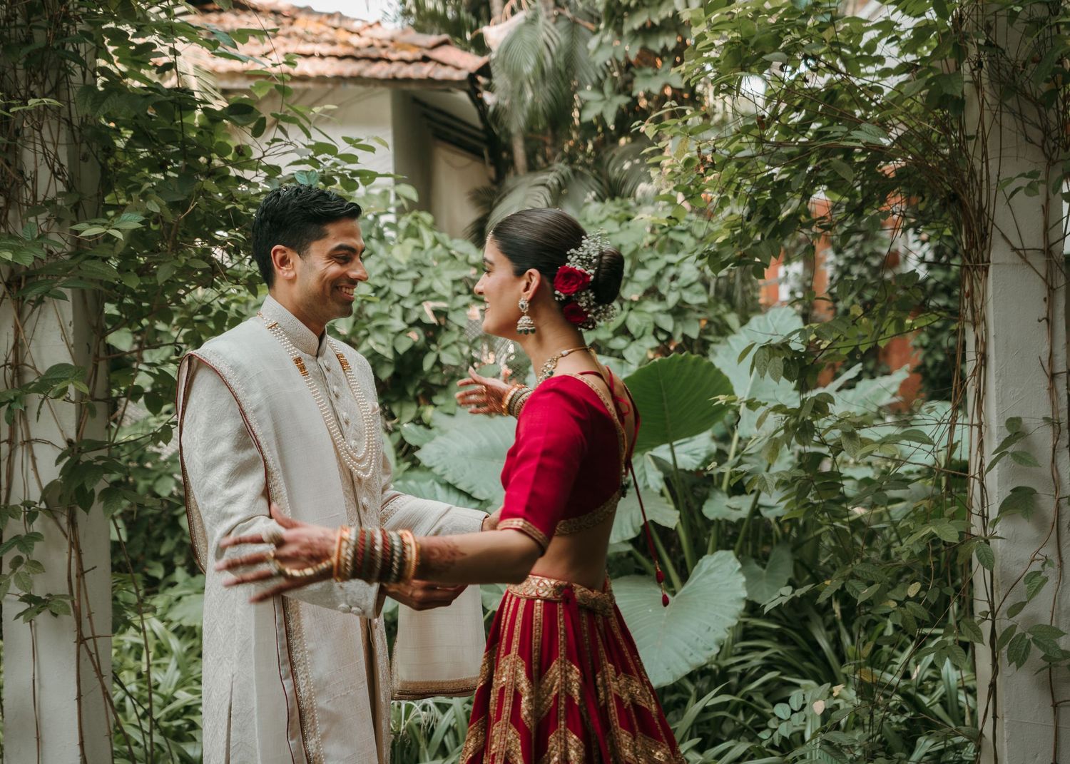 Indian couple in traditional wedding attire sharing a joyful moment surrounded by lush green foliage.