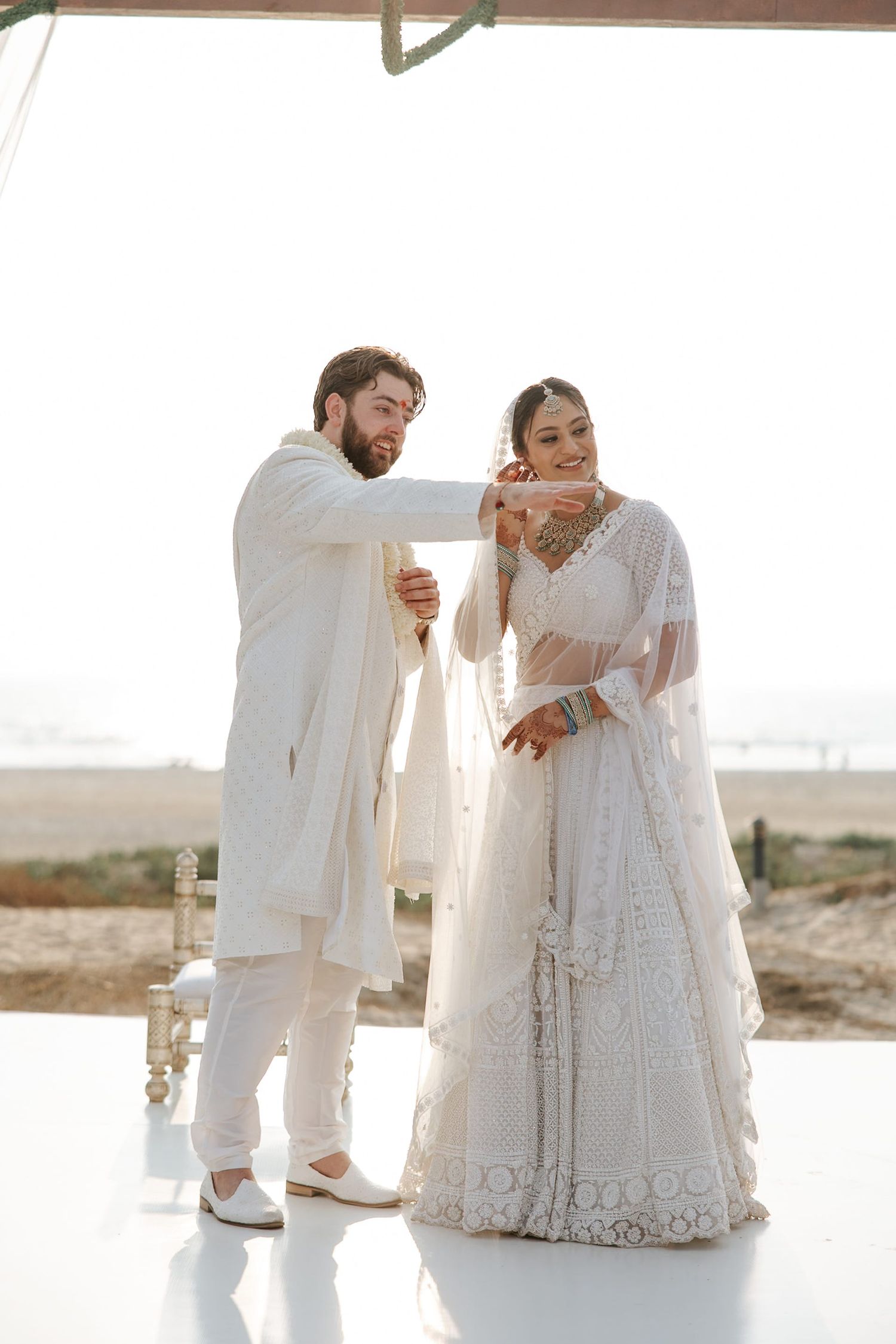 Couple in matching white Indian wedding attire, groom in kurta and bride in embroidered lehenga, sharing a tender moment outdoors.