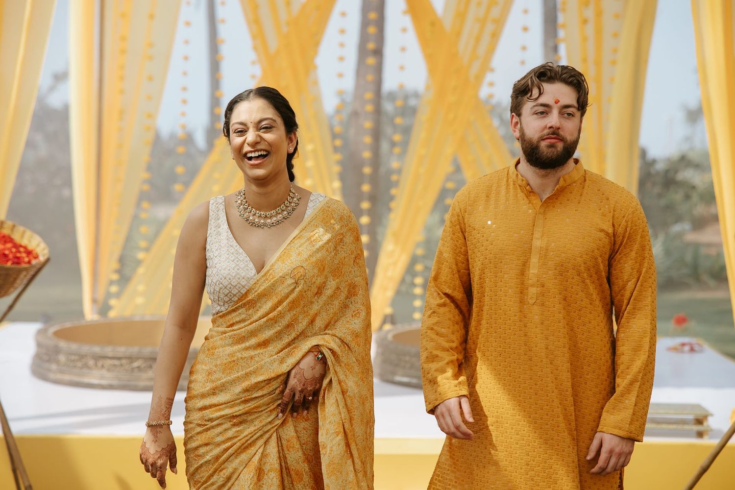 Couple in matching mustard yellow Indian wedding attire smiling at an outdoor ceremony.