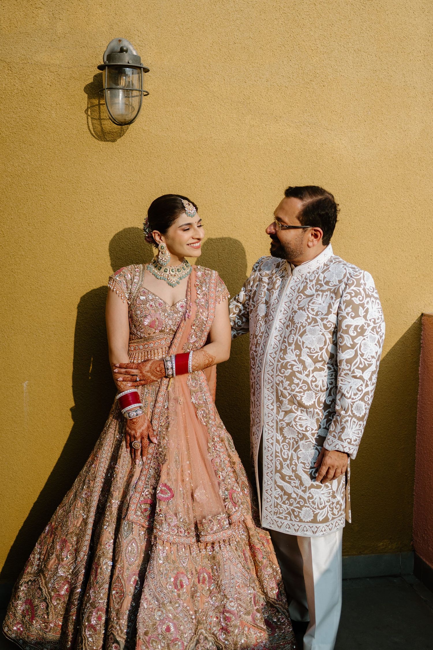 A couple in traditional South Asian wedding attire share intimate moments against a golden wall with vintage lamps.