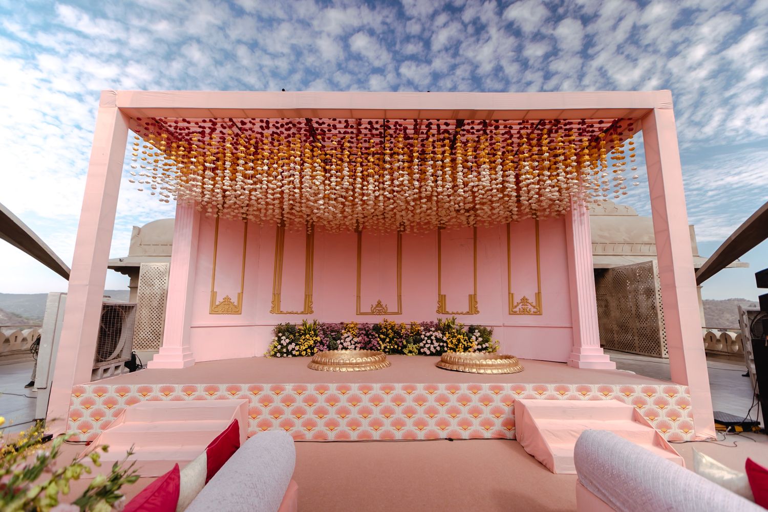 Pink outdoor wedding mandap with hanging floral chandelier decorations and white seating against a cloudy sky.