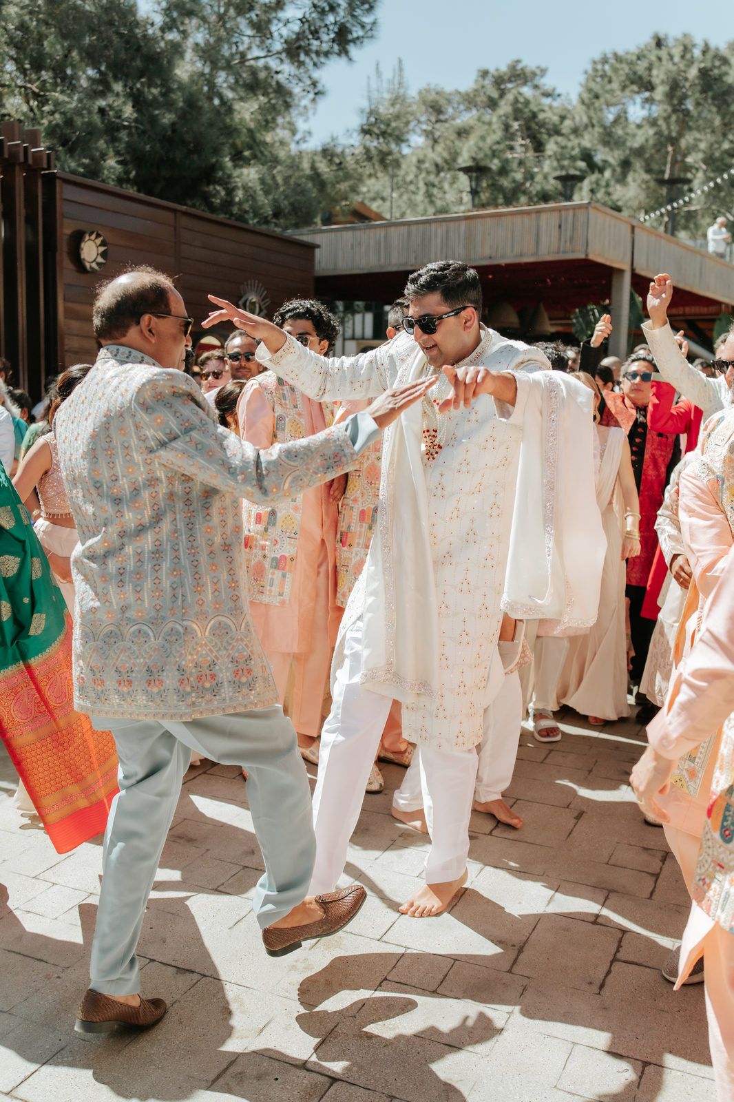Man in white kurta dances with elder during a vibrant outdoor Indian wedding celebration.