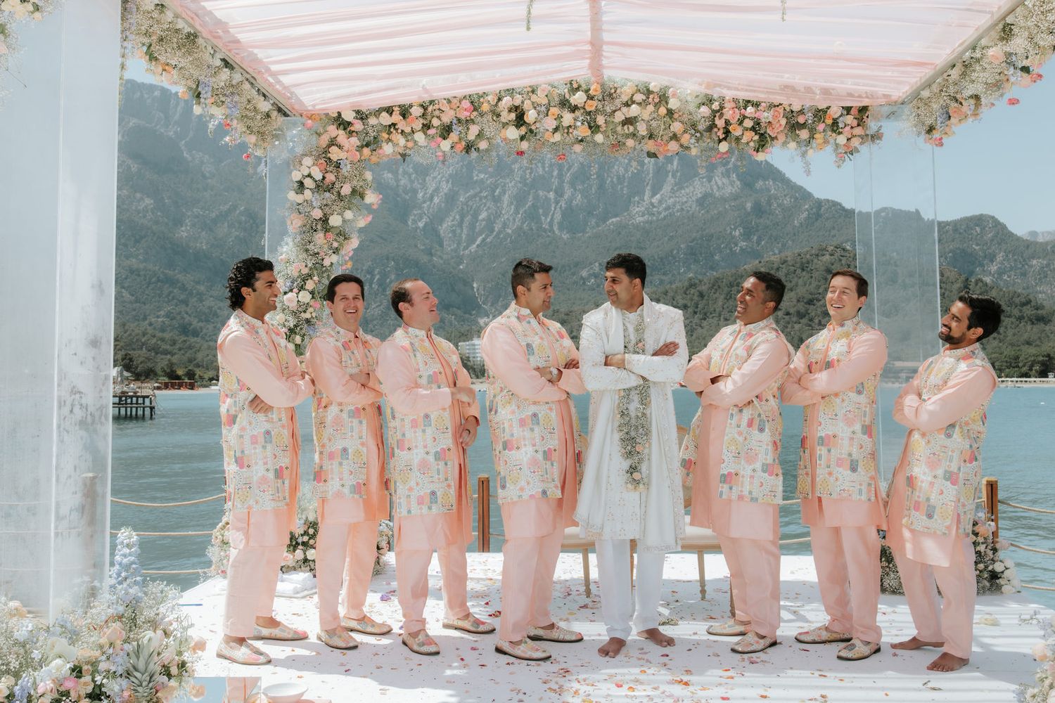 Groom in white sherwani poses with groomsmen in floral pink suits under decorated mandap by mountain lake.