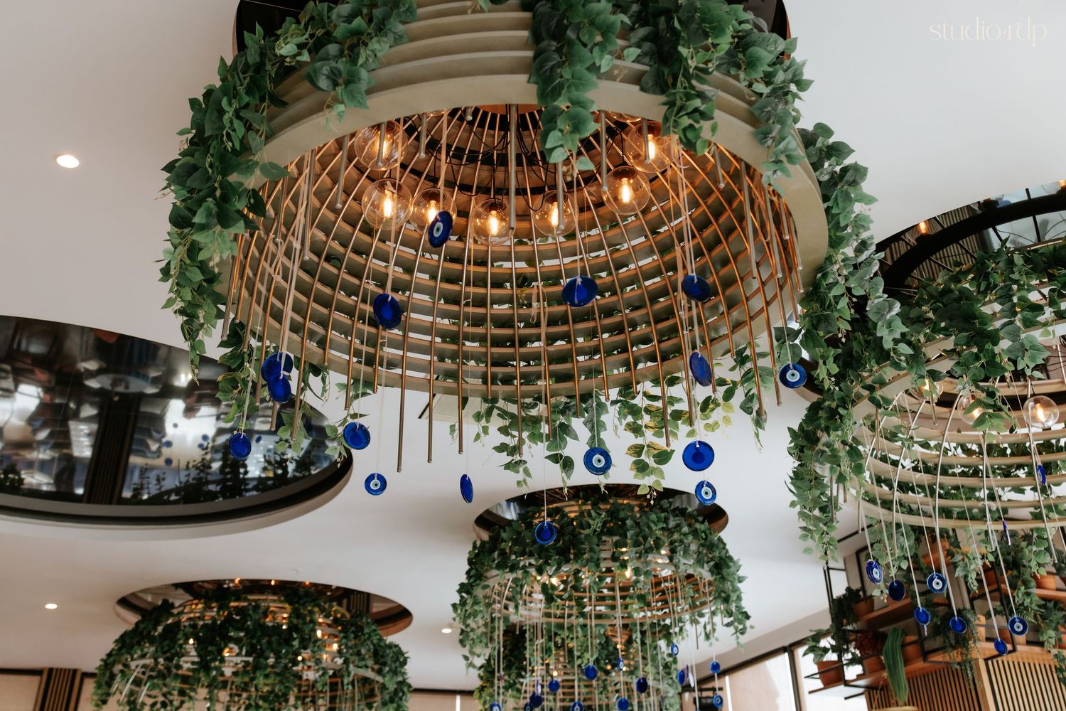 Ornate hanging chandeliers adorned with lush greenery, warm Edison bulbs, and blue evil eye charms in a stylish restaurant.