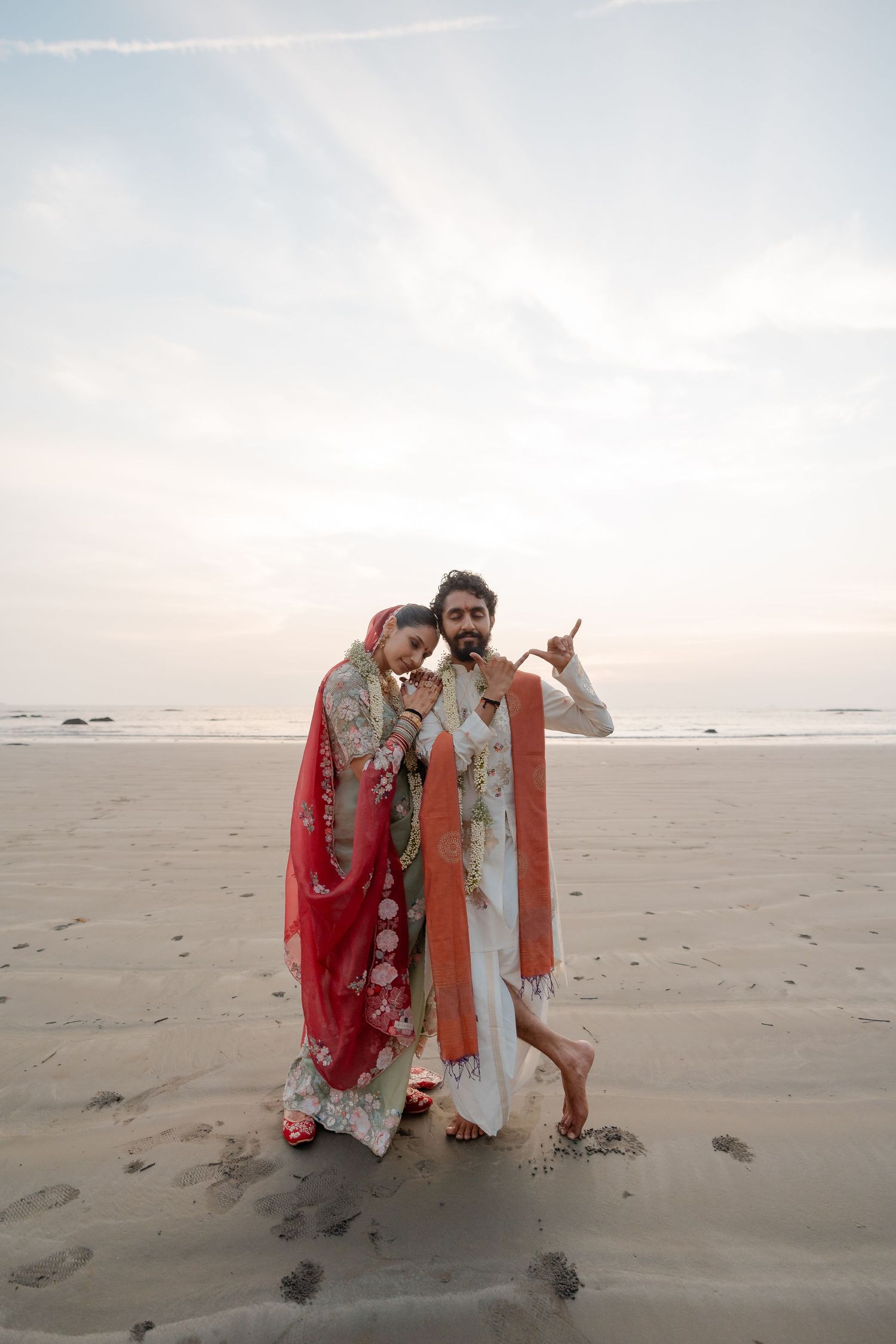 Couple in traditional South Asian attire posing on a sandy beach at dusk with ocean in background.