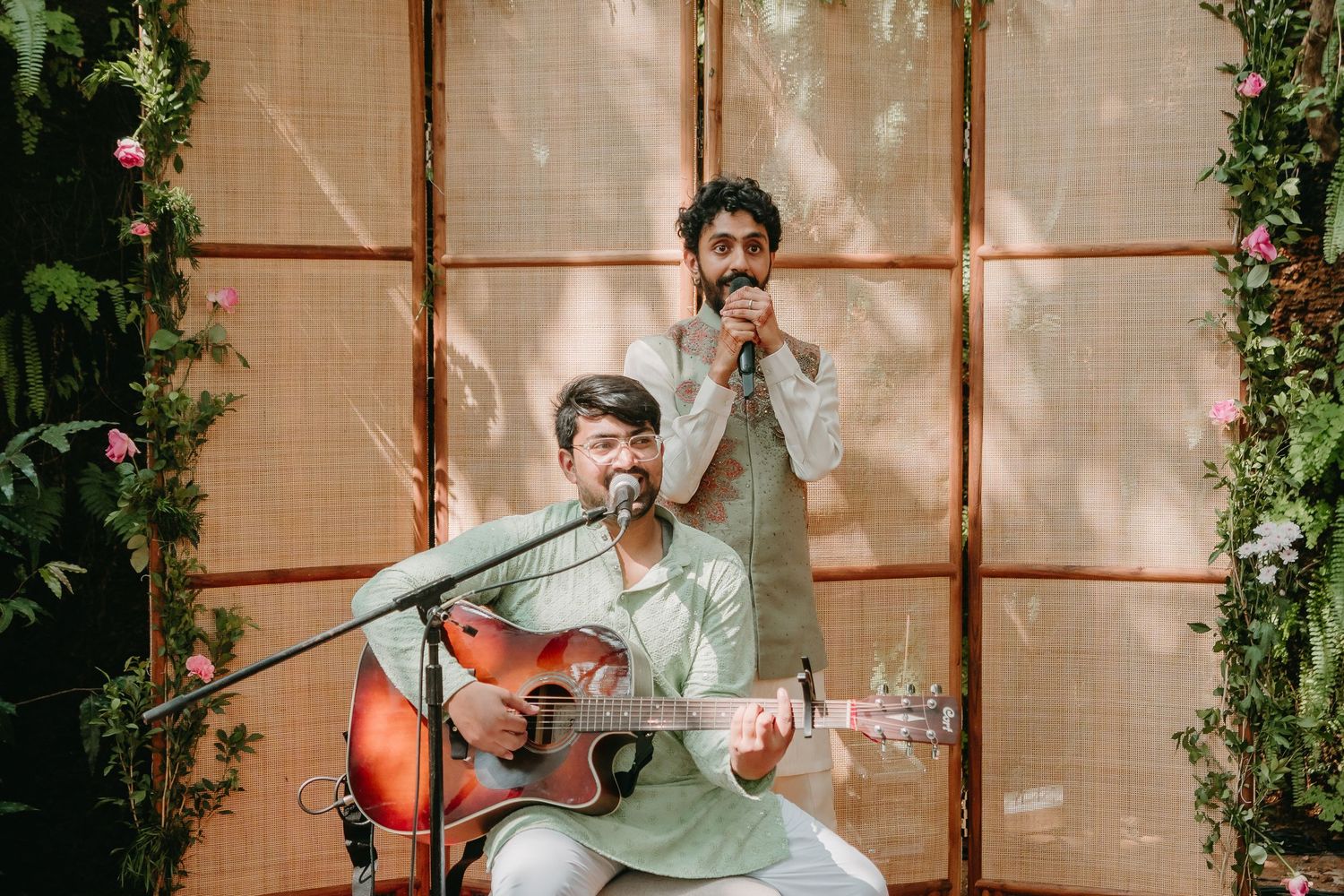 Two musicians perform outdoors, one playing acoustic guitar while the other sings into a microphone, surrounded by lush greenery.