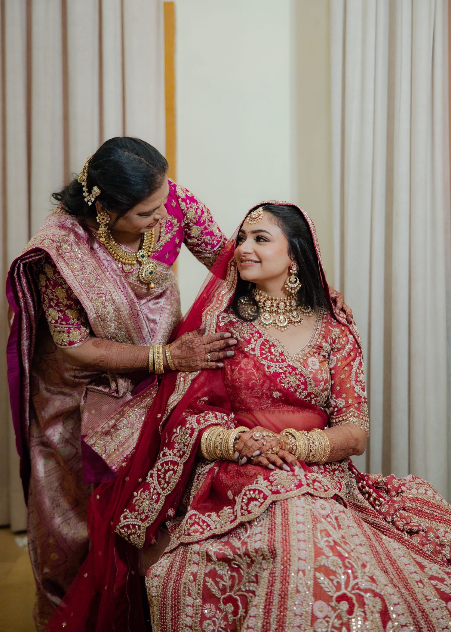 A bride in red lehenga with gold jewelry smiles as a woman in pink saree adjusts her dupatta during Indian wedding.