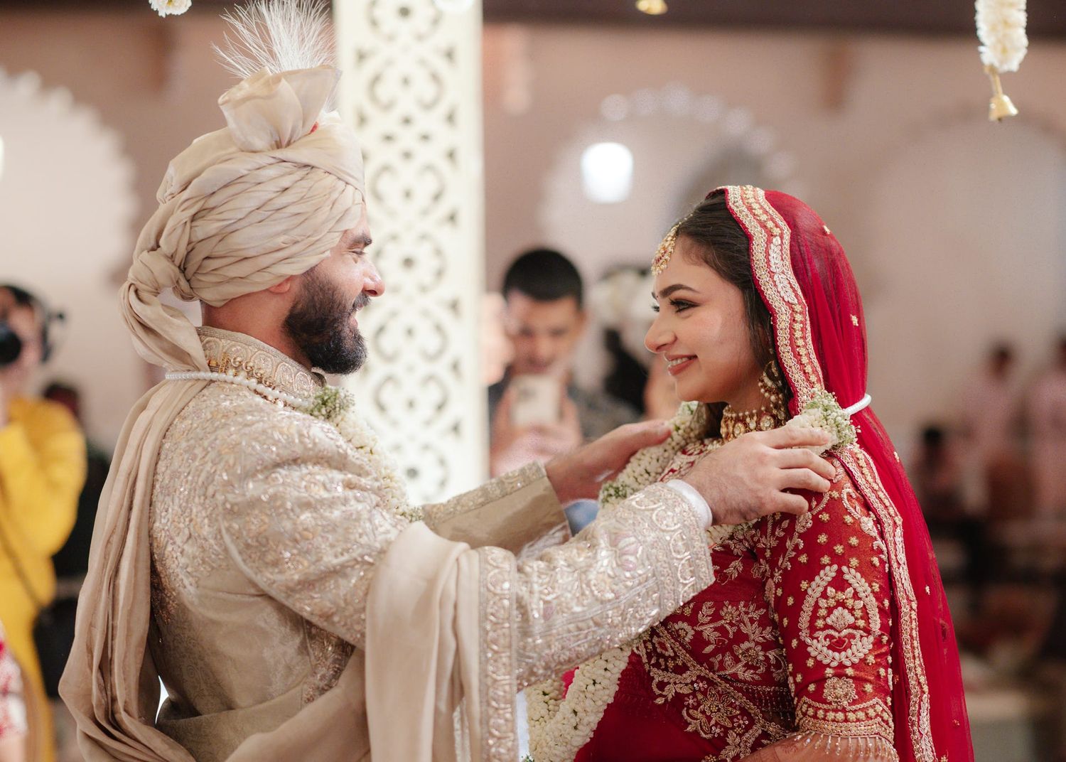 Bride in red lehenga and groom in cream sherwani exchange garlands at traditional Indian wedding.