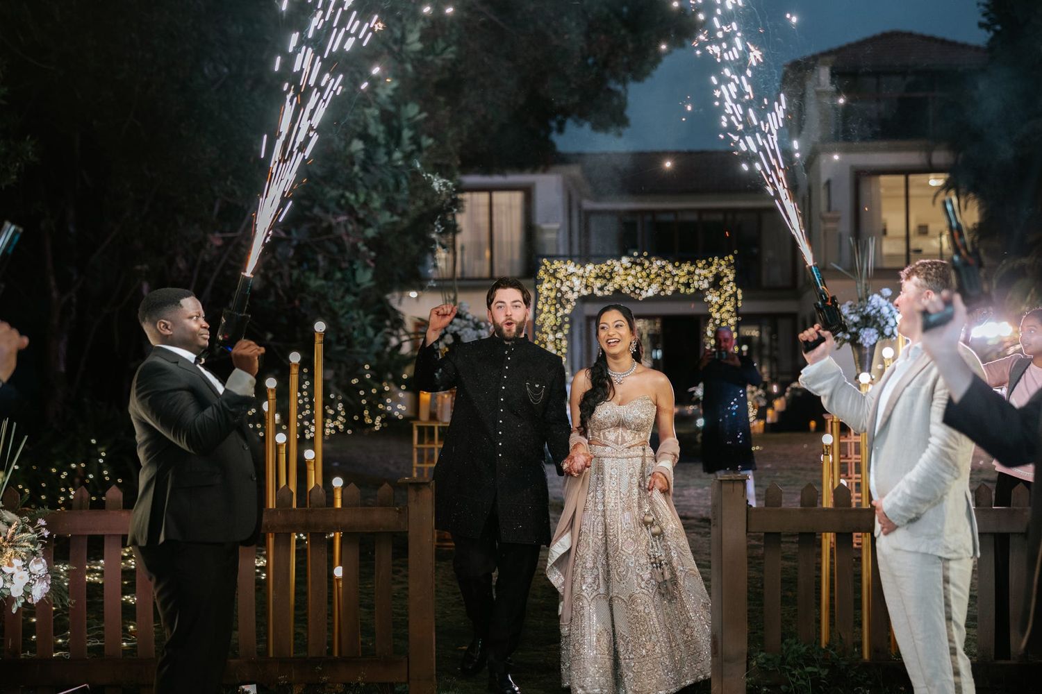 Bride and groom exit wedding ceremony through cold spark fountains at elegant outdoor night reception.