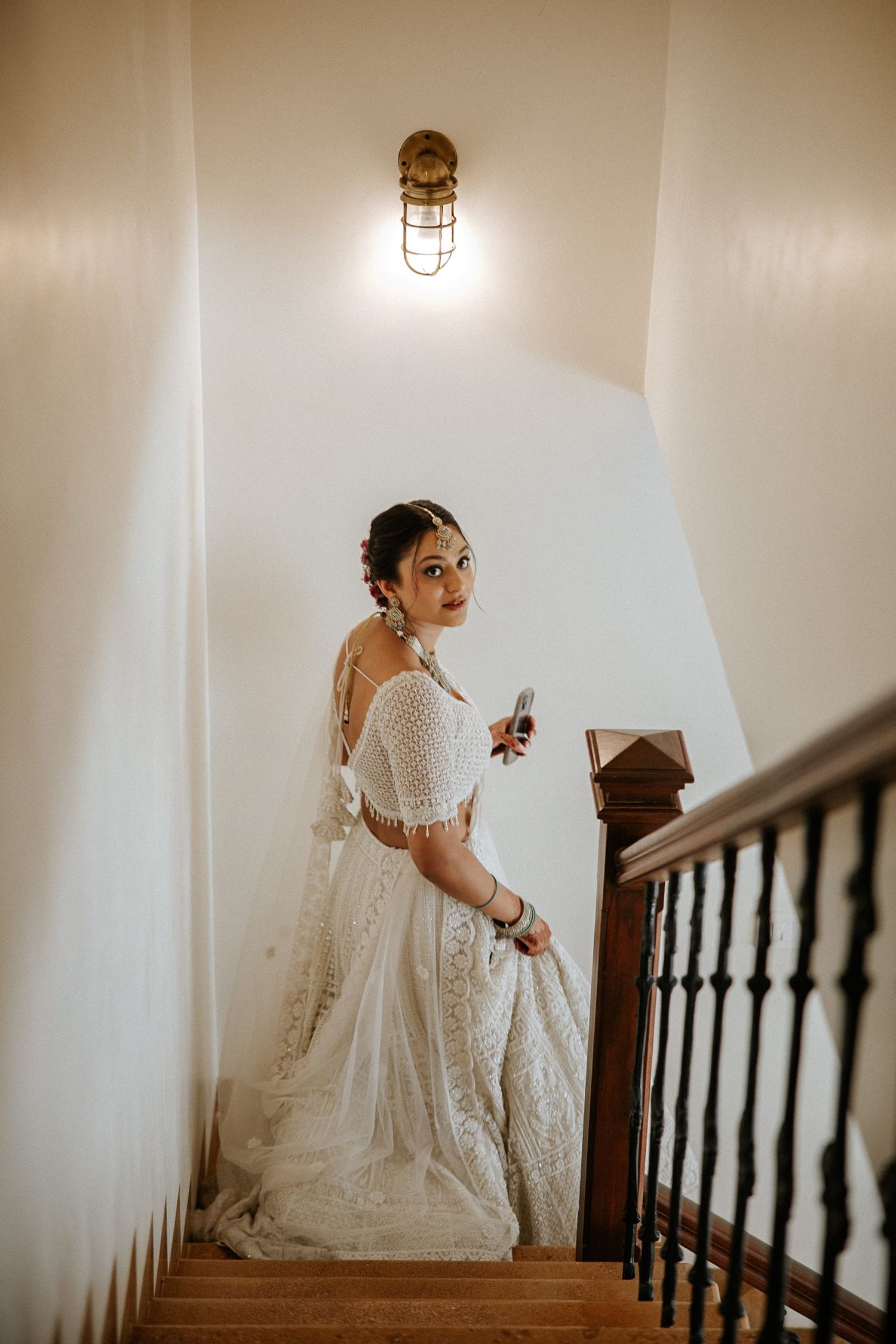 Bride in white embroidered lehenga stands on staircase holding phone, adorned with jewelry and dupatta, soft warm lighting.