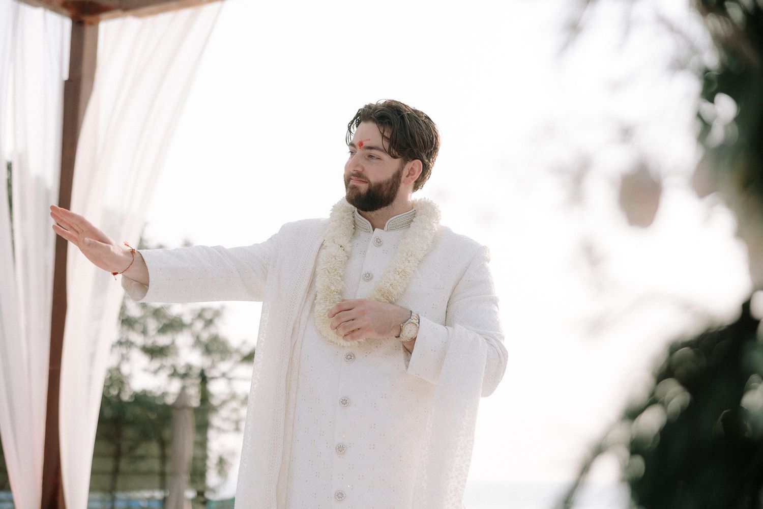 Groom in white sherwani with floral garland gestures during outdoor Indian wedding ceremony.