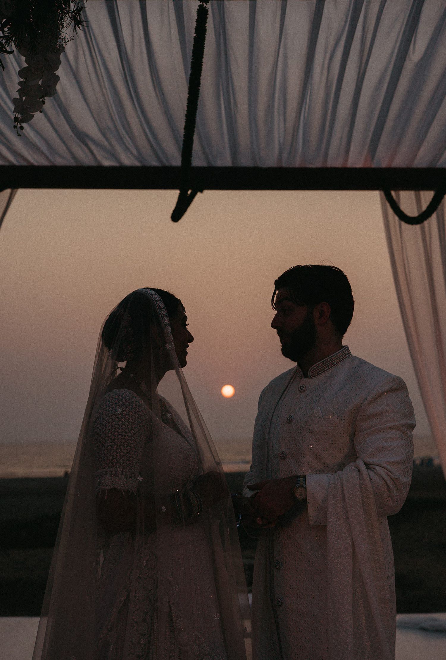 Couple in traditional Indian wedding attire silhouetted against a stunning sunset backdrop under a draped canopy.