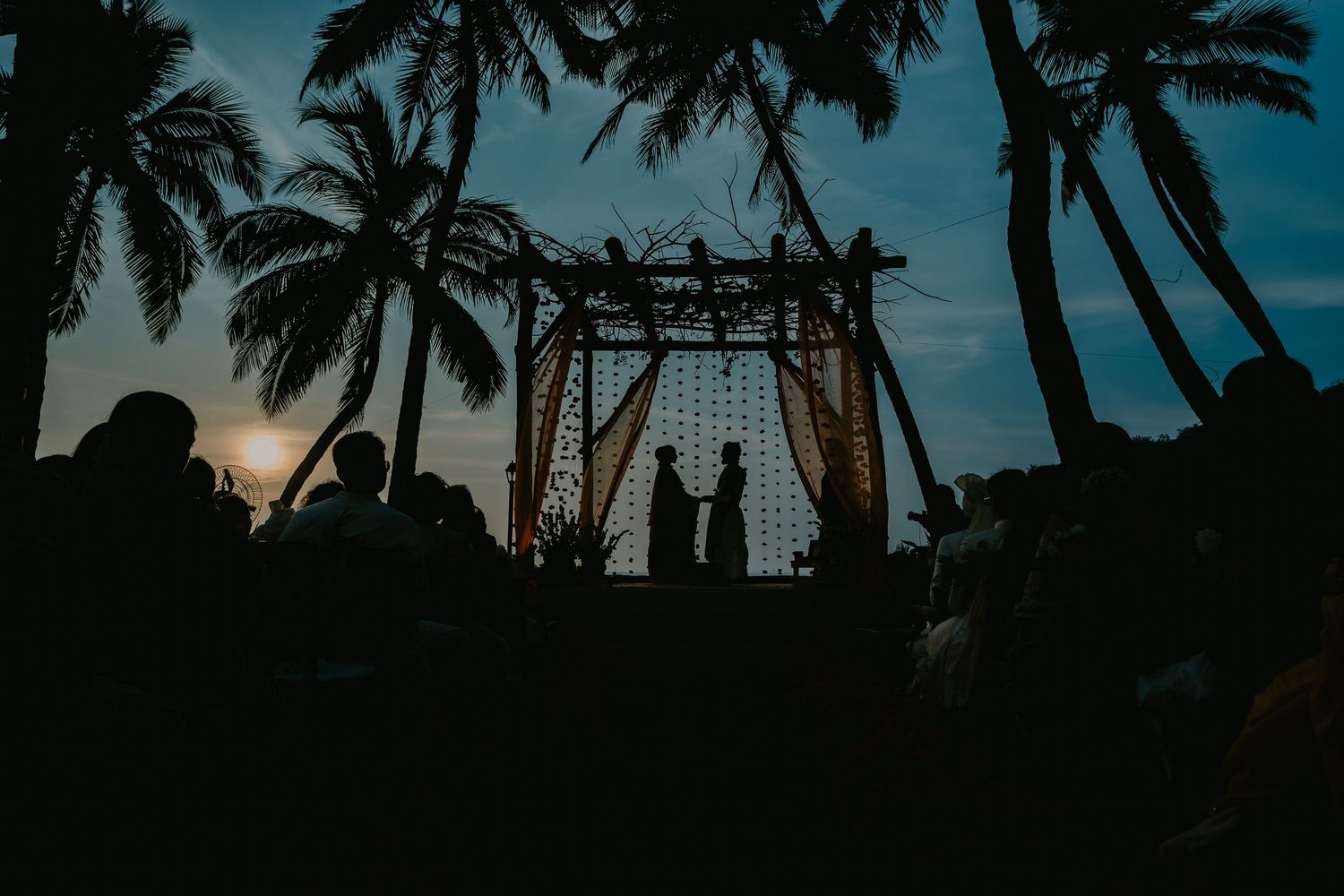 Silhouette of couple at beach wedding ceremony under palm trees at sunset with guests seated.
