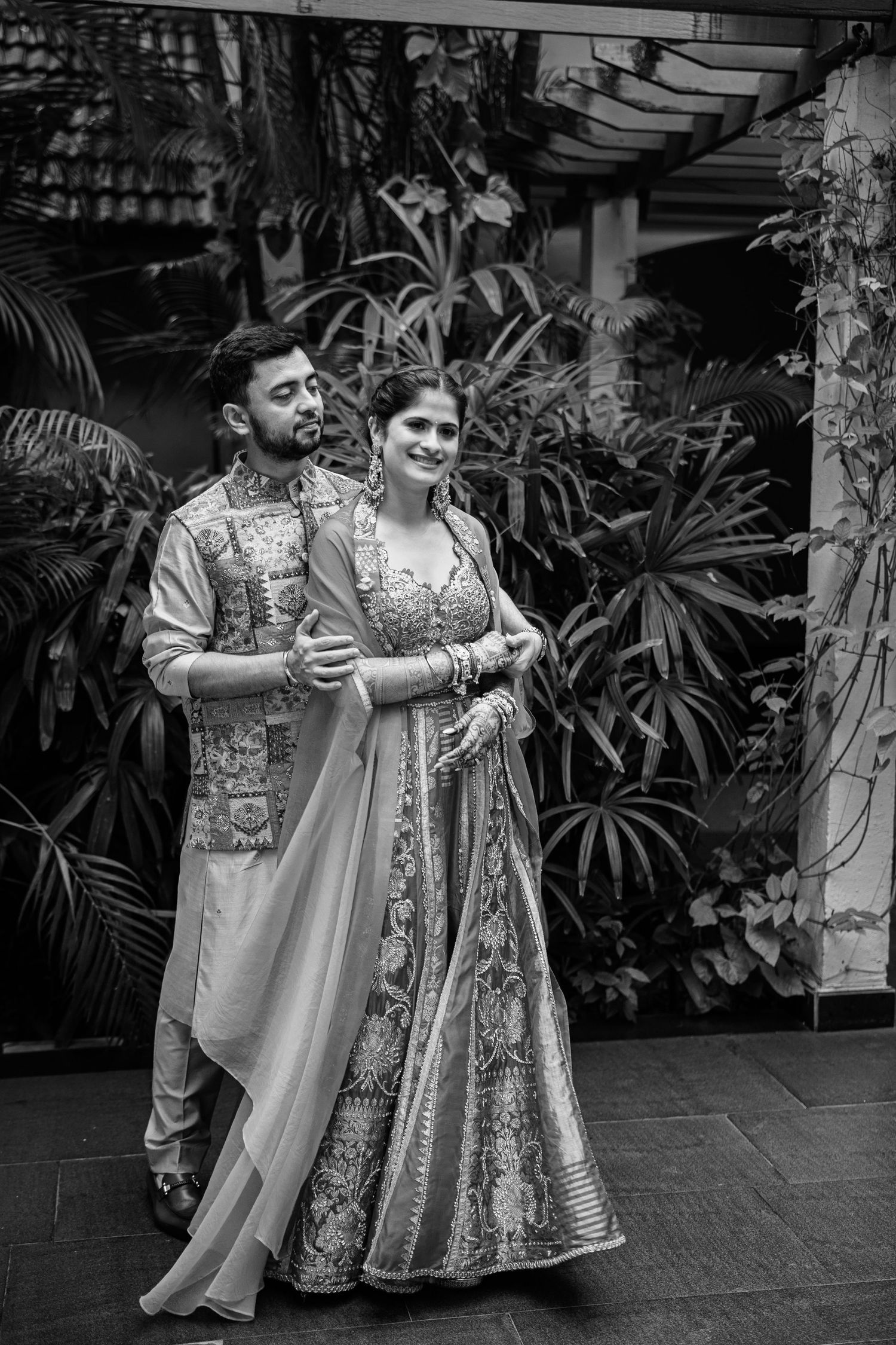 Black and white photo of an Indian couple in traditional wedding attire posing together among lush tropical greenery.