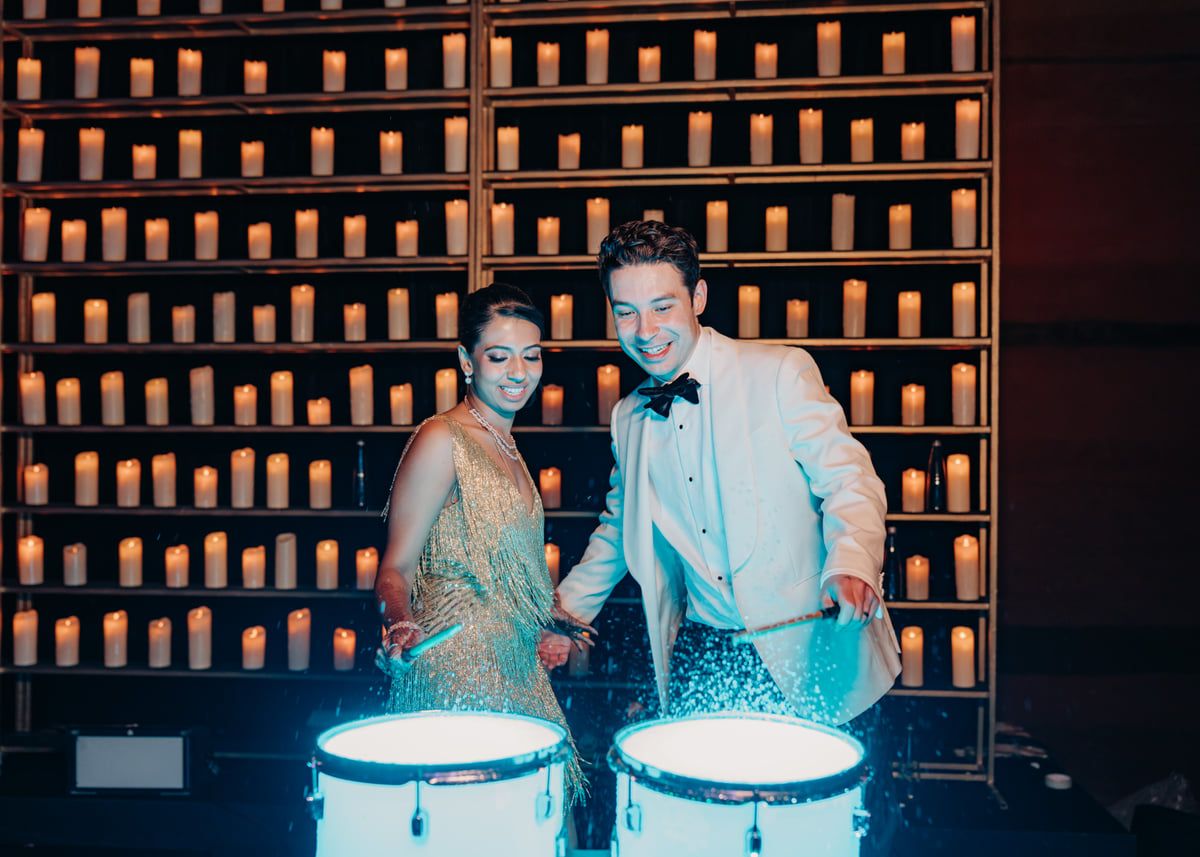A couple stands in formal wear against a backdrop of wooden shelves with illuminated bottles.