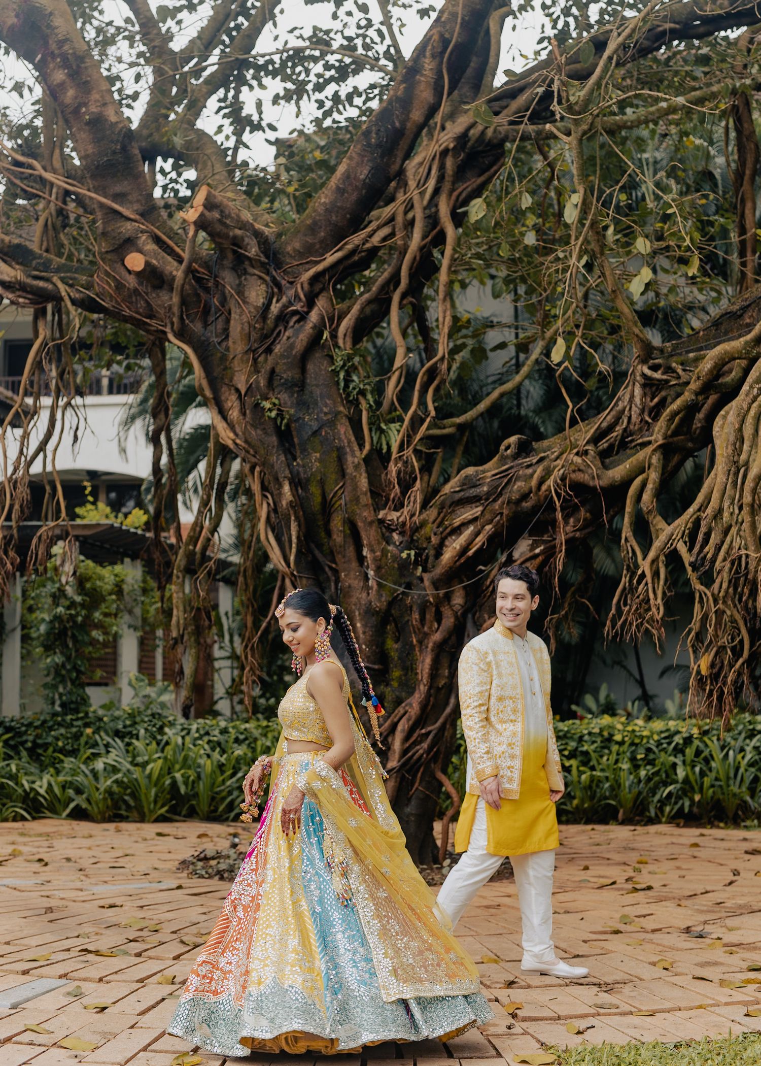 Couple in vibrant Indian wedding attire walking before a sprawling banyan tree, bride in colorful lehenga, groom in yellow kurta.