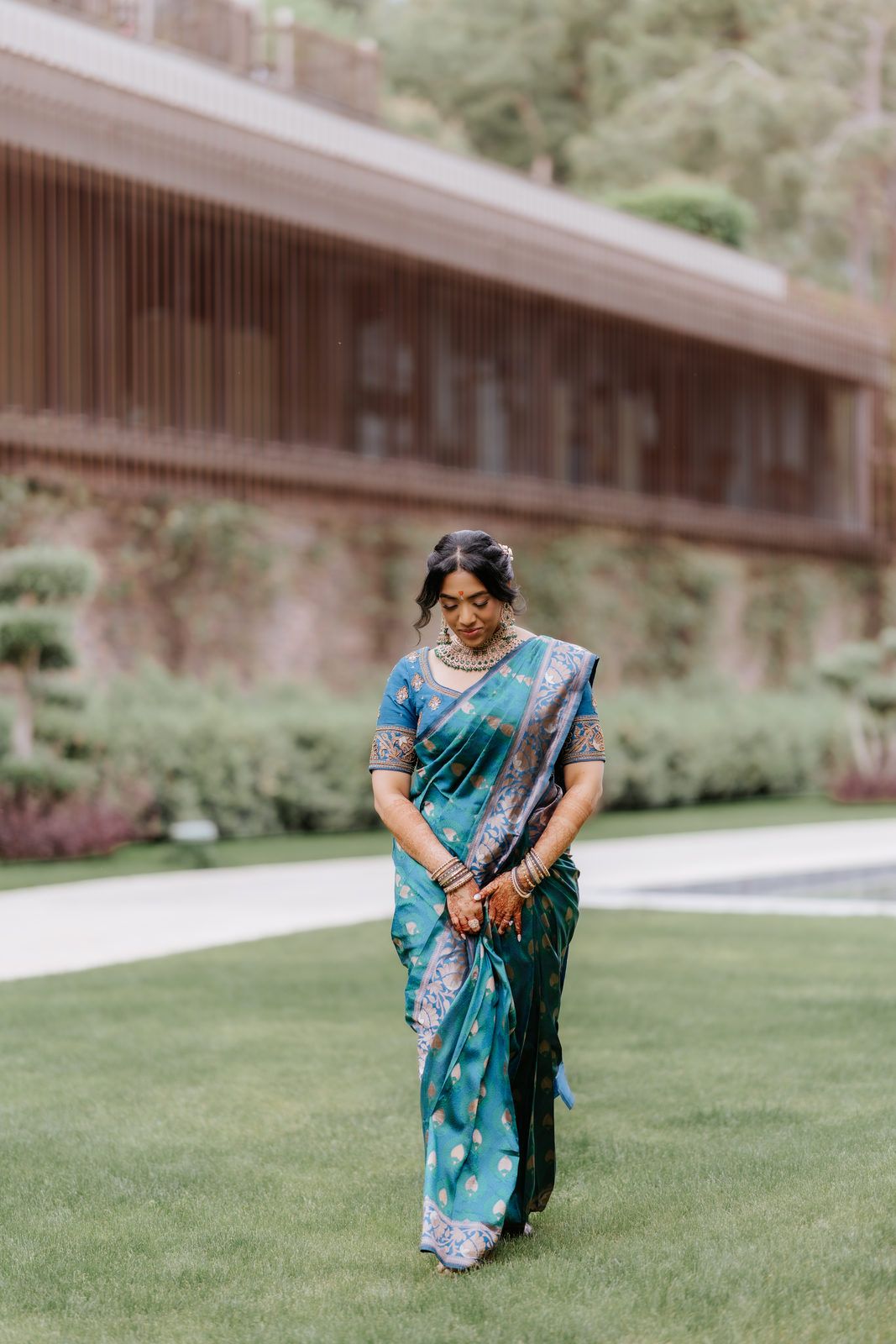 Woman in teal silk saree with gold embroidery walking gracefully on green lawn at elegant outdoor venue.
