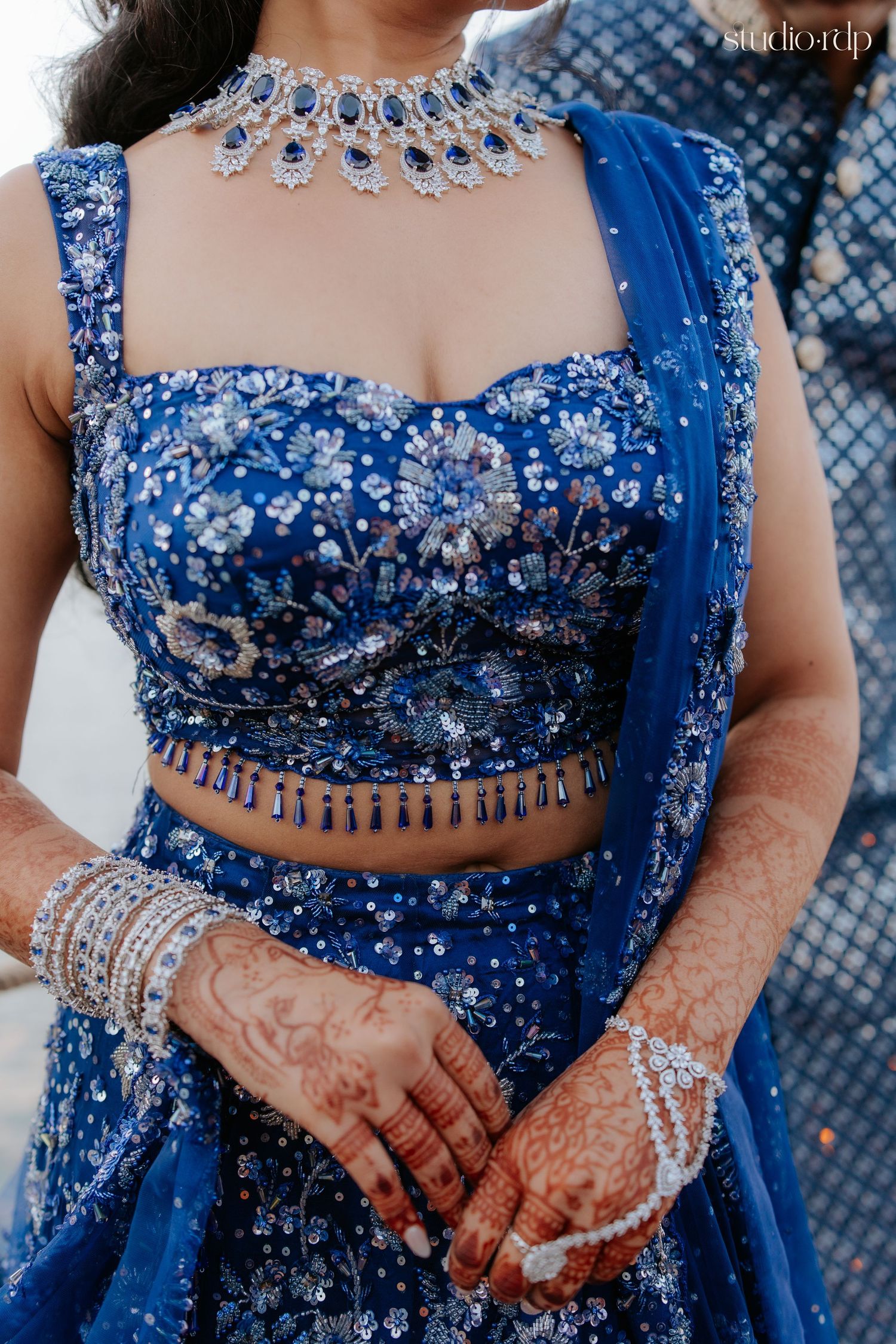 Bride in royal blue embroidered lehenga with floral motifs, diamond necklace, bangles, and intricate mehndi on hands.